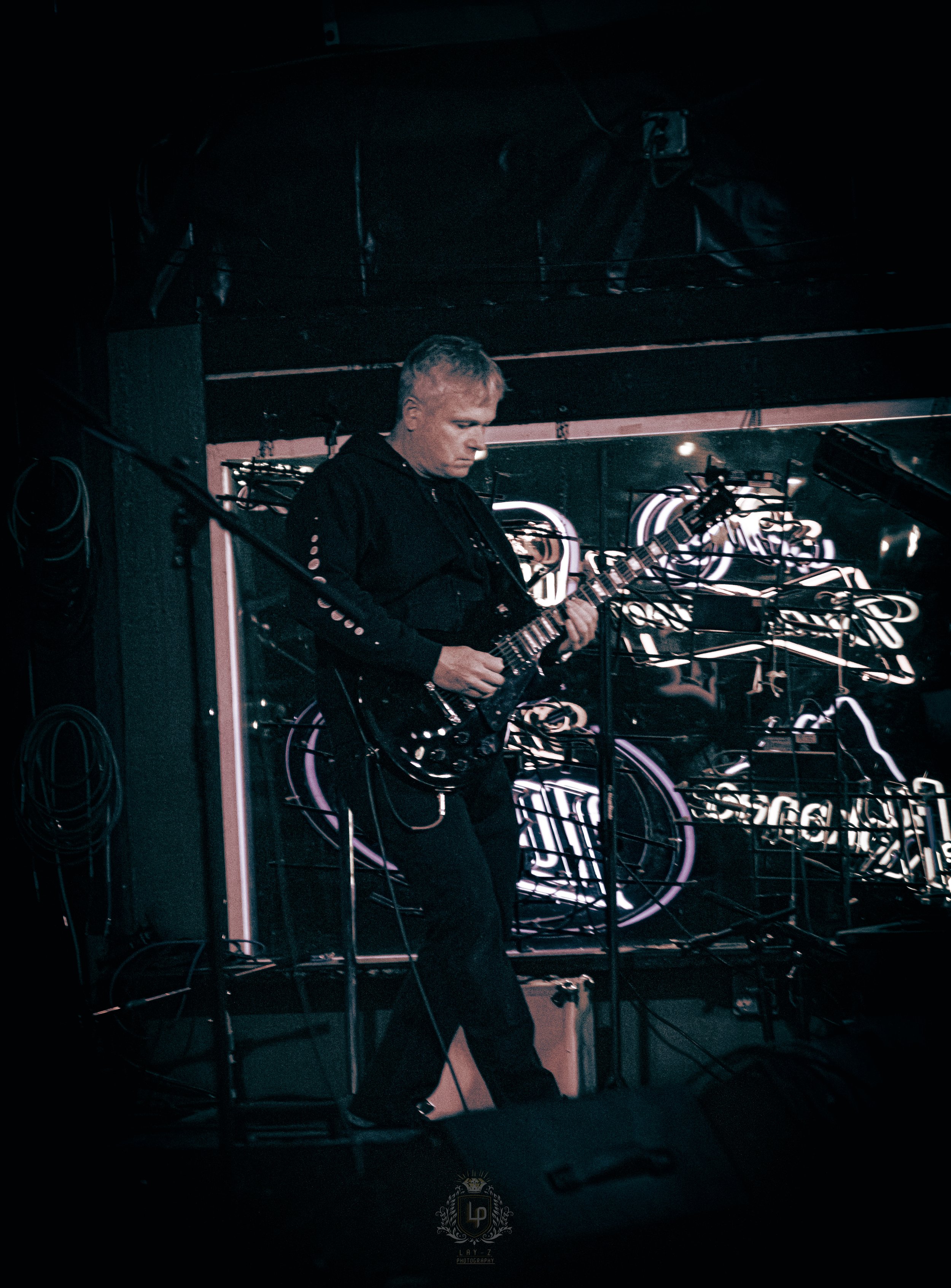 A man playing an electric guitar on stage with a neon sign in the background.