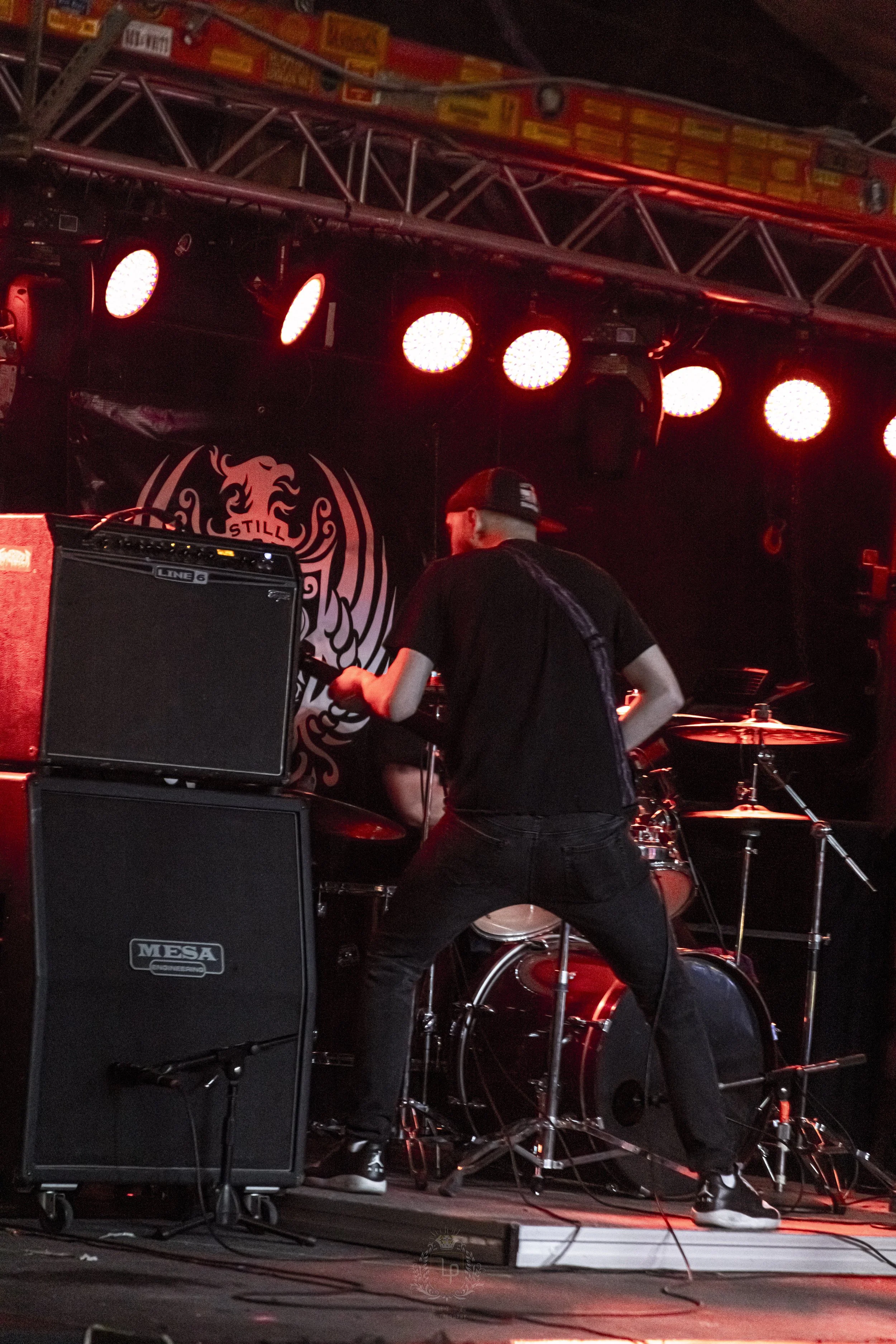 A musician on stage preparing to play drums, with amplifiers and stage lighting visible, during a live music performance.
