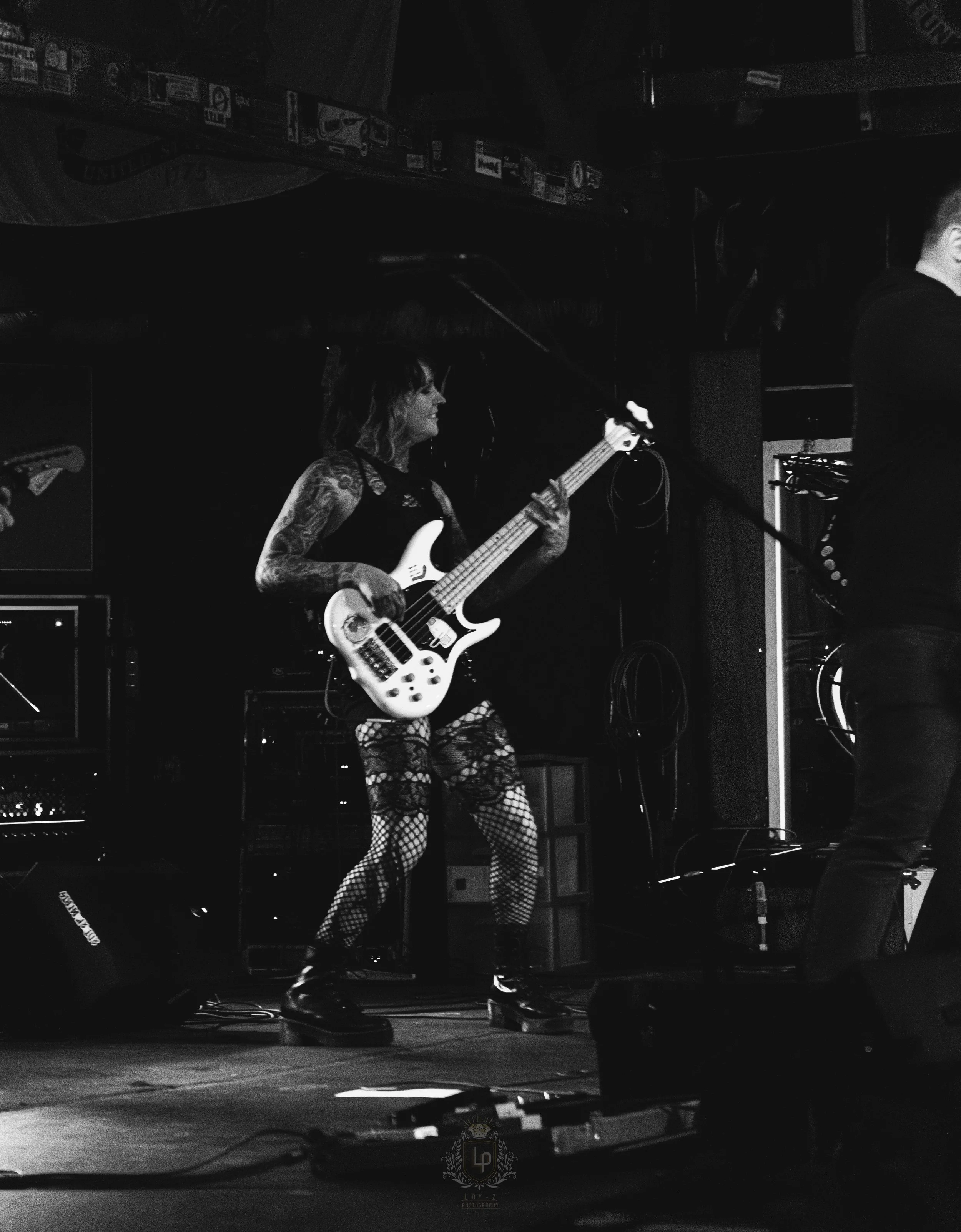 A female musician playing a bass guitar on stage, wearing fishnet stockings and lace clothing, with tattoos on her arms, in a dark venue.