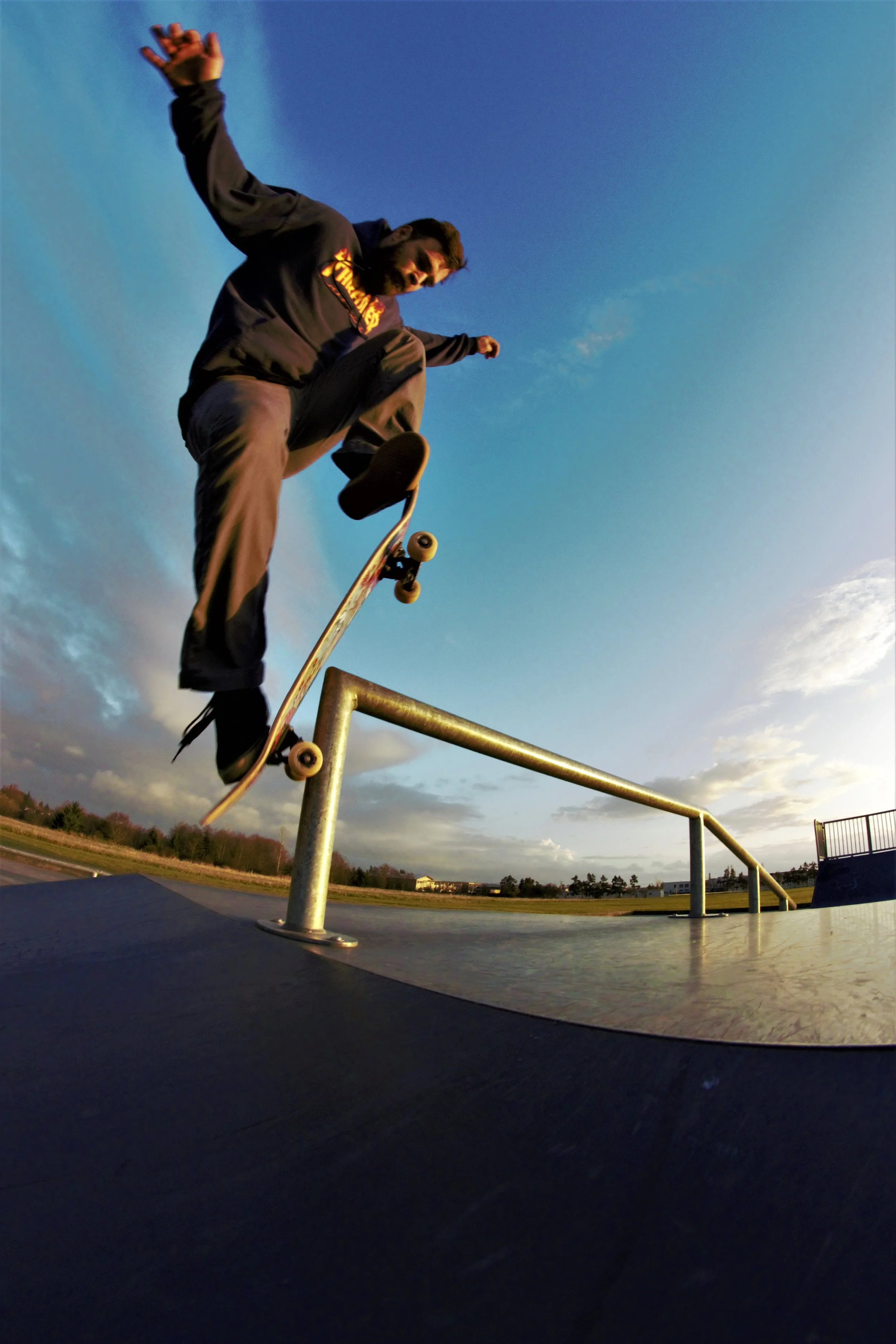 Skateboarder performing a trick on a rail at a skate park with a sunset sky in the background.