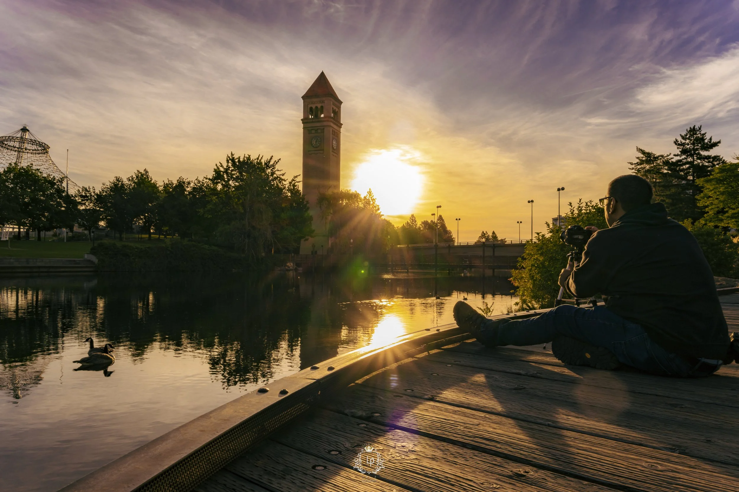 Person sitting on a wooden dock by a body of water, taking a photo at sunset with a camera. Ducks swimming in the water, trees lining the shore, a clock tower in the background, and the sun setting behind the tower.
