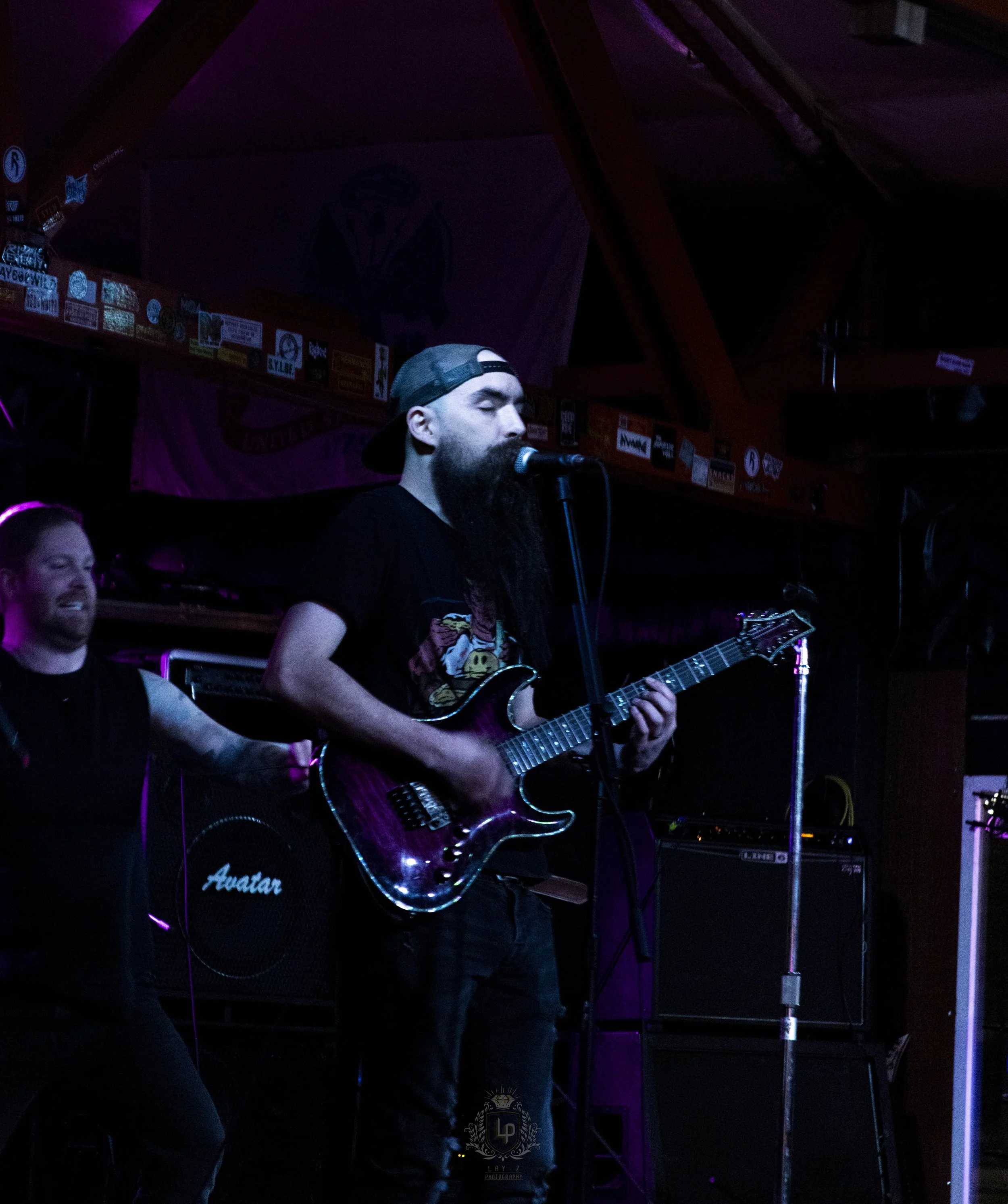 A man with a beard, wearing a backwards cap and a black t-shirt with a graphic, playing an electric guitar and singing into a microphone on stage. Another person is partially visible next to him, smiling. The background includes amplifiers and stage 