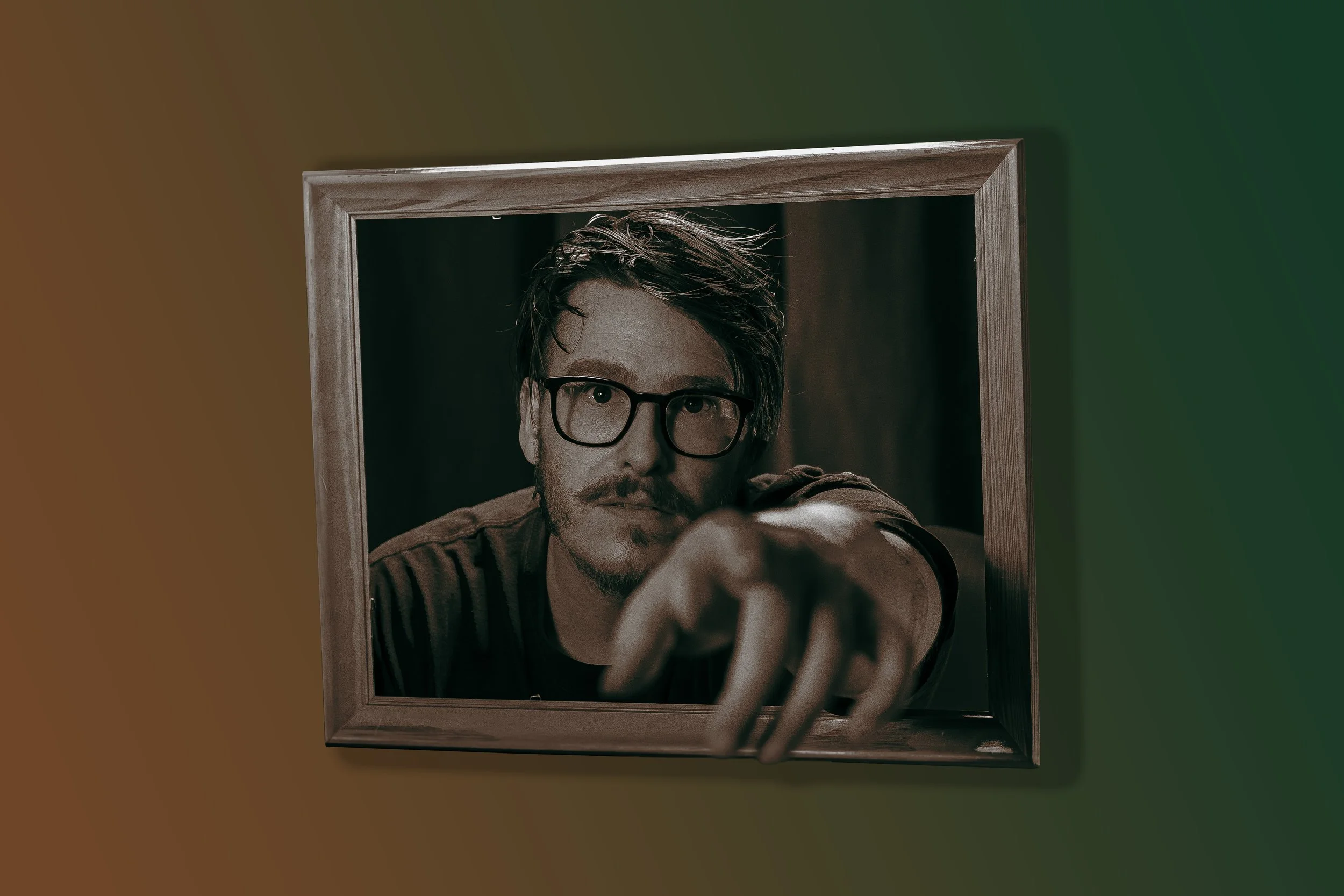A man with glasses and a mustache points towards the camera while looking through a wooden frame set against a multicolored background.