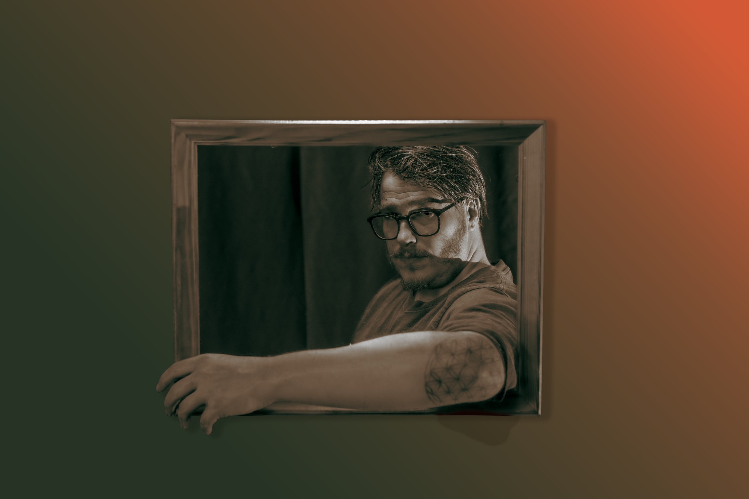 A man with glasses and styled hair holding a picture frame in front of himself, with a tattoo on his forearm visible. The background is a blurred gradient of warm colors.