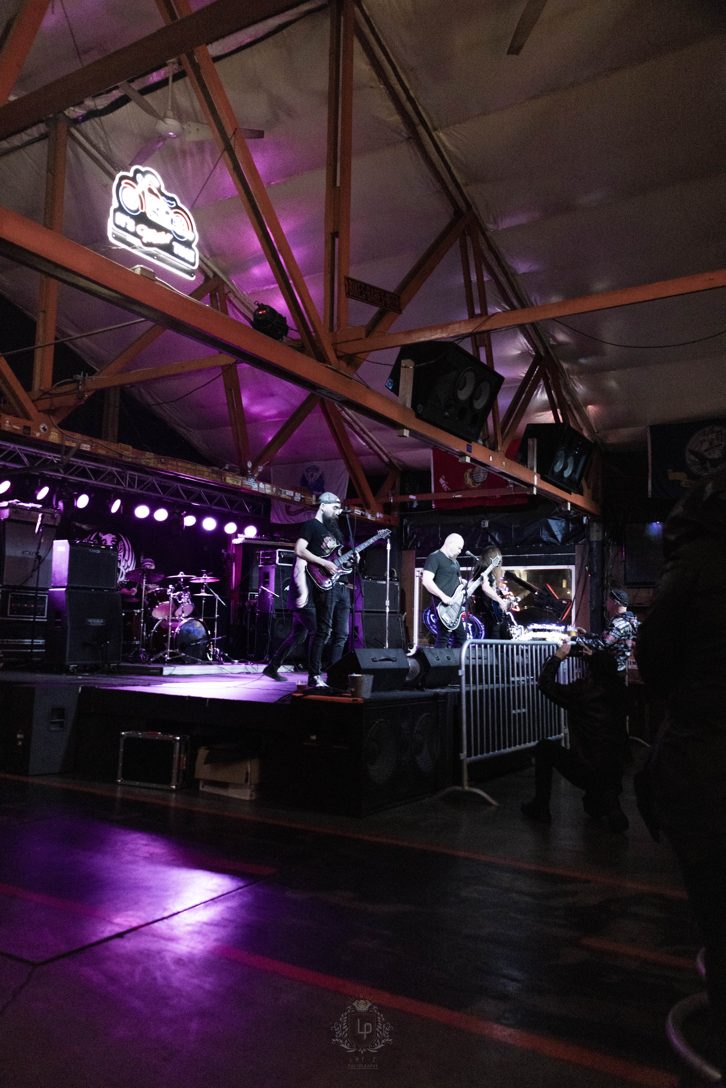 A live band performs on a stage with purple and pink lighting inside a venue, with a drummer, guitarists, and a vocalist visible. A photographer is taking pictures in front of the stage. Neon sign and large black speakers are also visible.