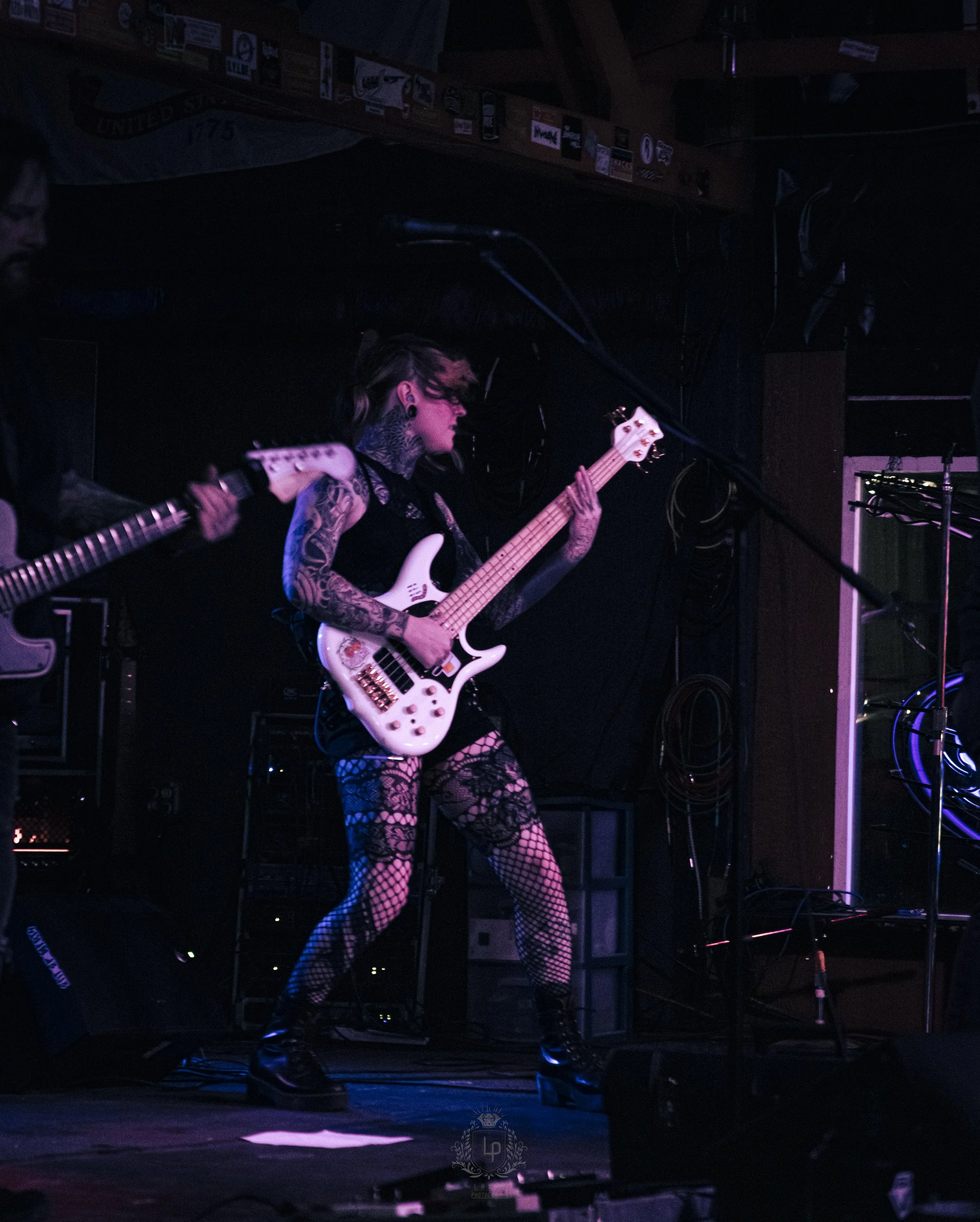 A woman with tattoos and fishnet stockings playing a white bass guitar on stage during a live music performance.
