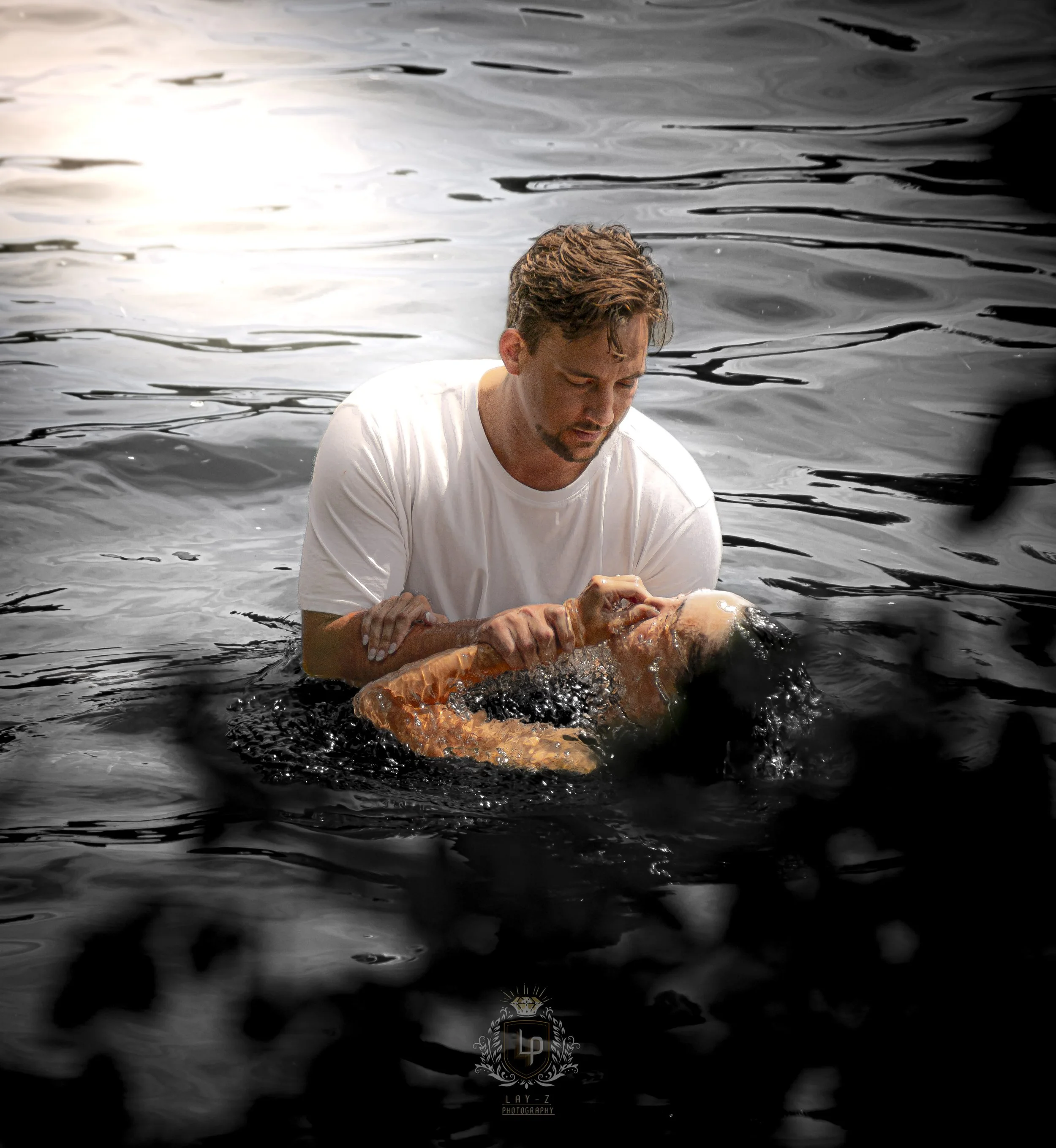 A man in a white shirt performing a rescue in a body of water, holding a woman in black with her eyes closed, surrounded by dark water.