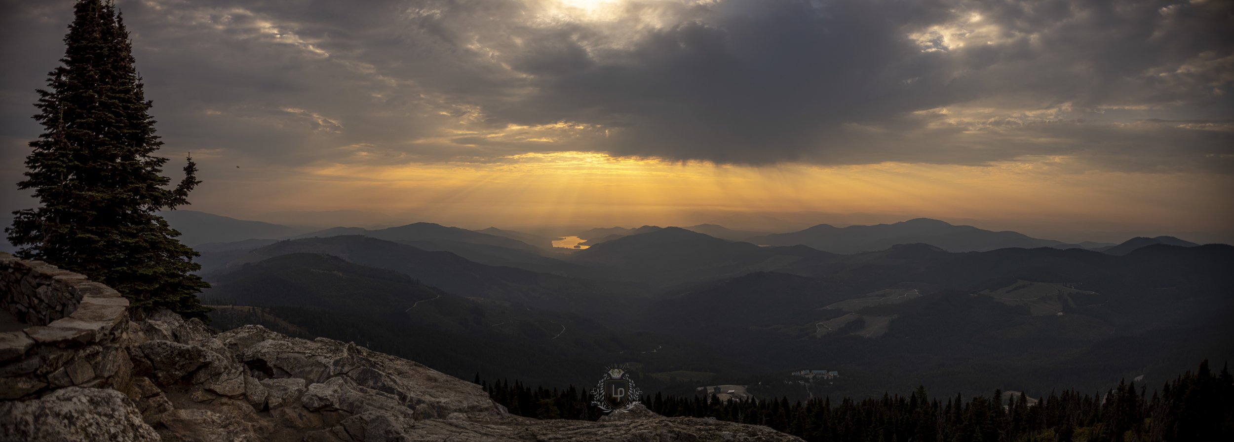 A scenic view of mountain ranges during sunset with clouds and rays of sunlight breaking through, and a lake in the distance.