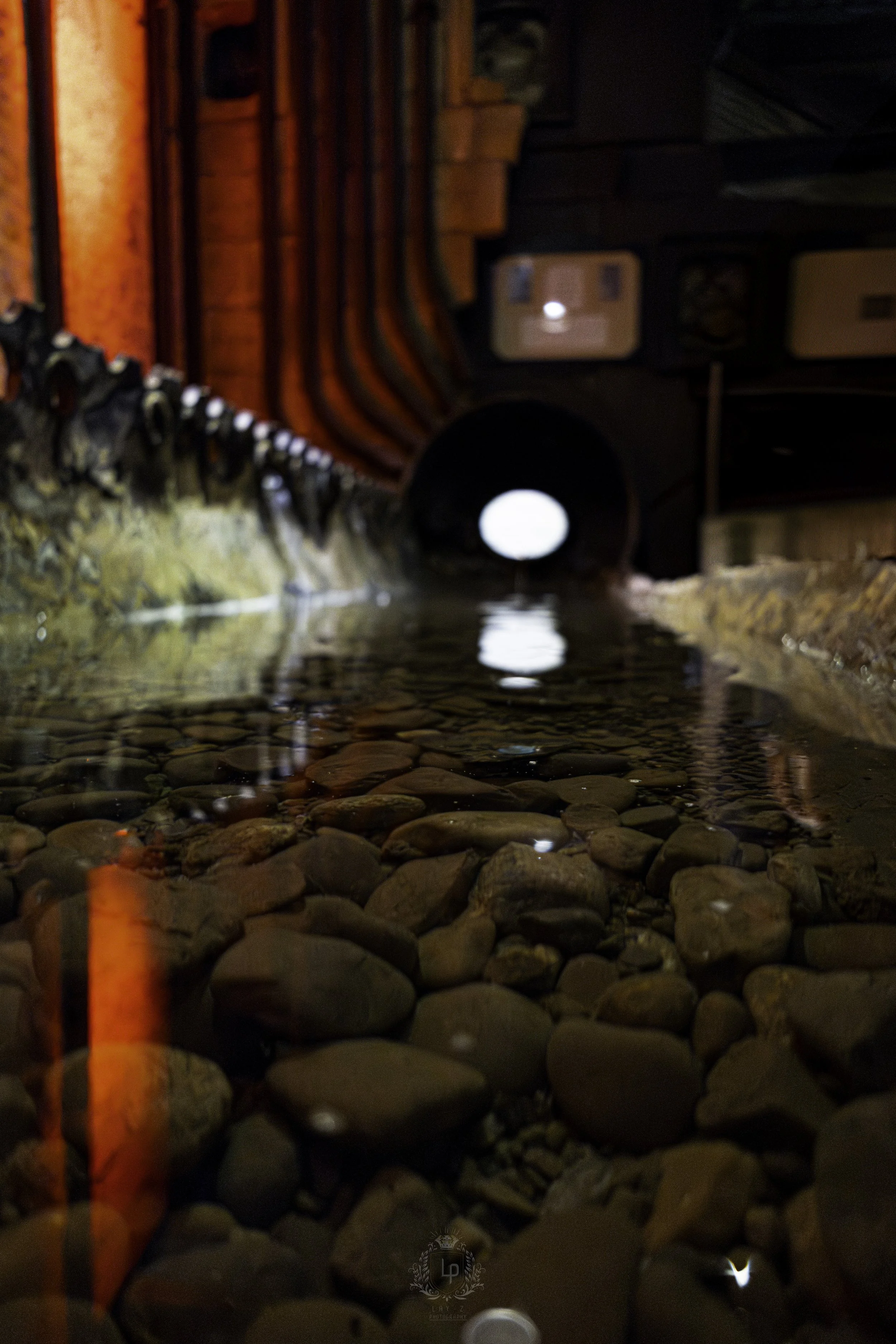 Close-up view of a rock-filled water channel with a circular drain pipe in the background and red-painted pipes on the left.