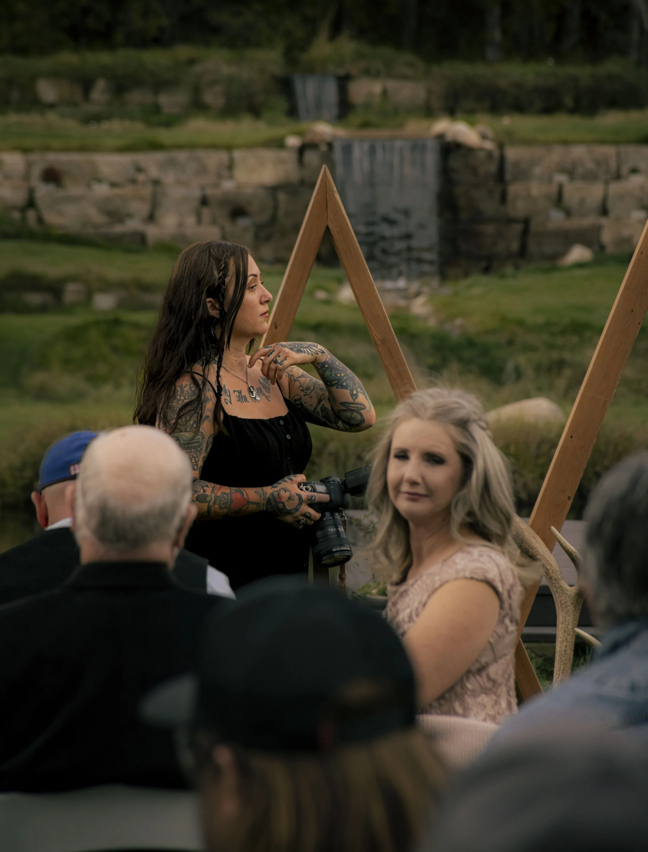 A woman with tattoos holding a camera talks during an outdoor event with seated audience, with a natural landscape and waterfall in the background.