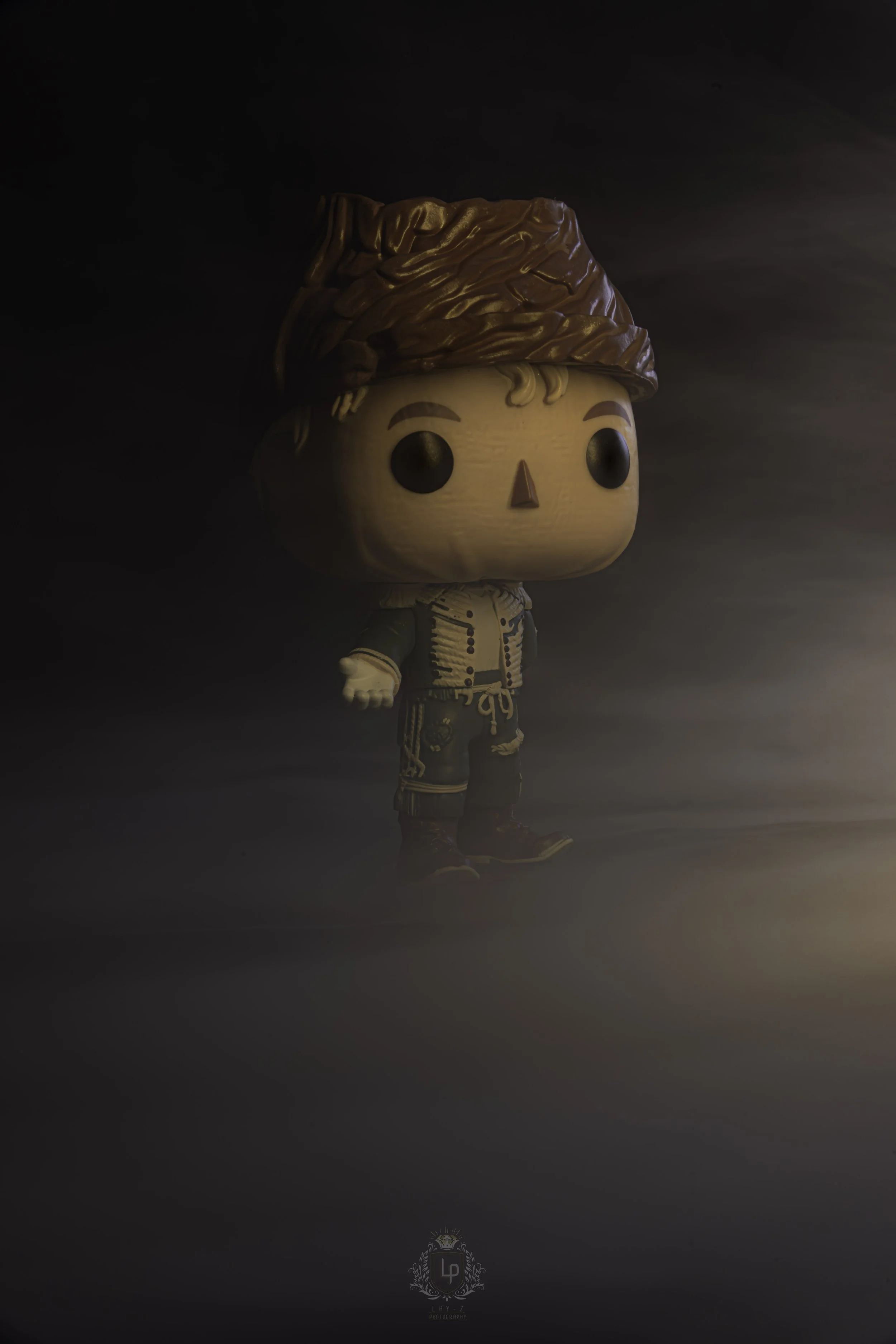 A Funko Pop! figure of a young man with curly blonde hair wearing a brown hat, green jacket, and dark pants, standing in a dark, foggy environment with a soft light illuminating him from the side.