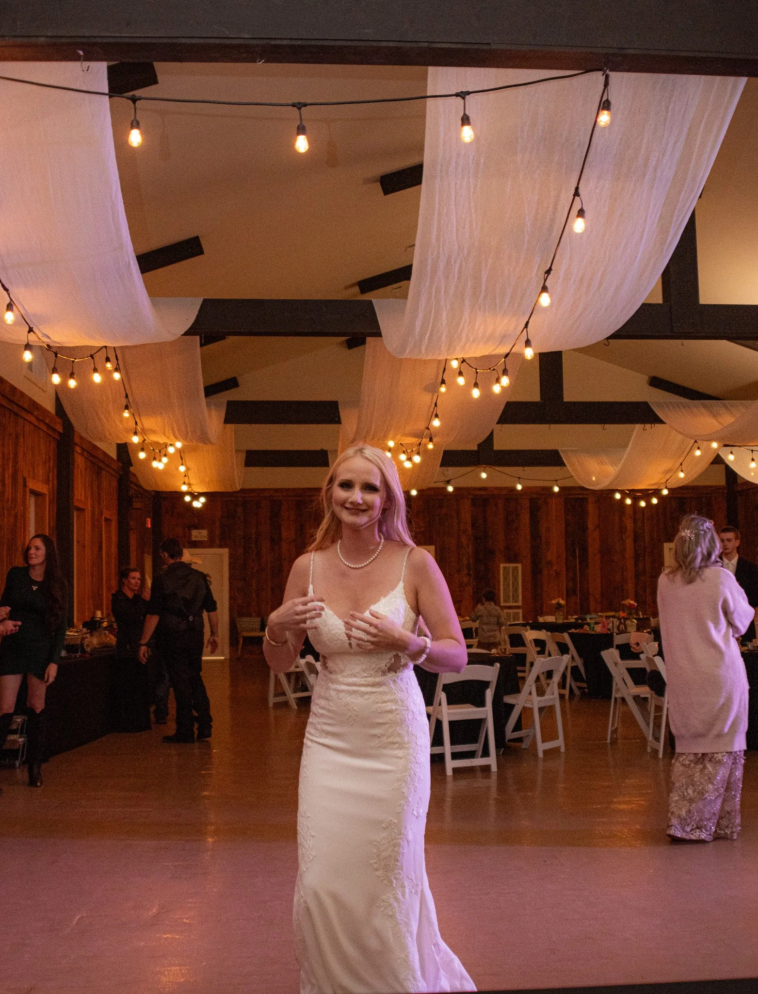 A woman in a white wedding dress smiling at the camera while standing on a dance floor at a wedding reception venue decorated with string lights and fabric drapes hanging from the ceiling.