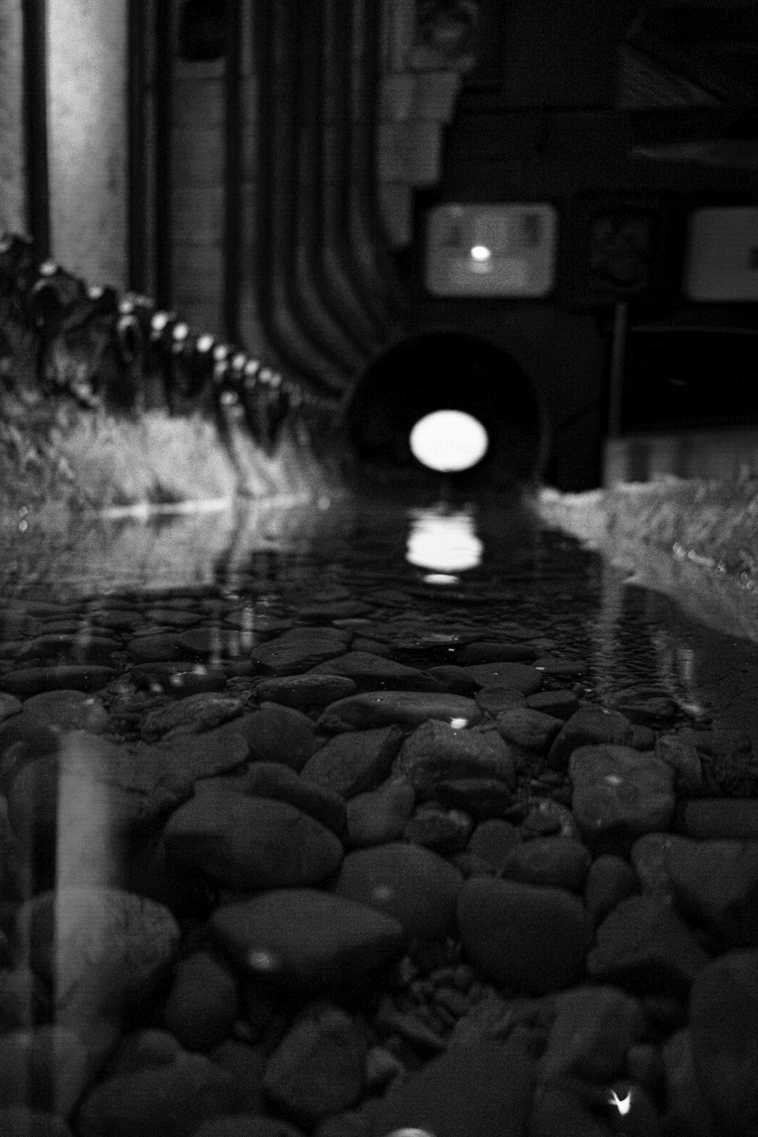 A black and white photo of a drainage tunnel with water flowing on a rocky bottom, seen from a low perspective at the water level, with a bright circular opening at the far end.