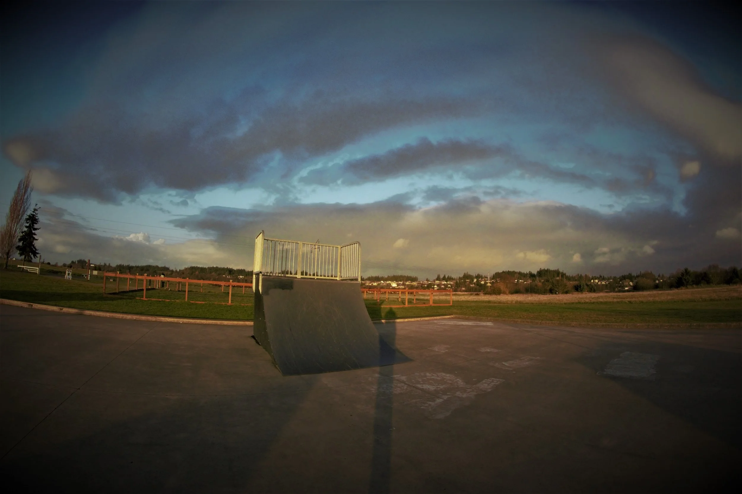 A skateboarding ramp at a skate park with cloudy sky, trees, a grassy field, and distant houses and trees in the background.