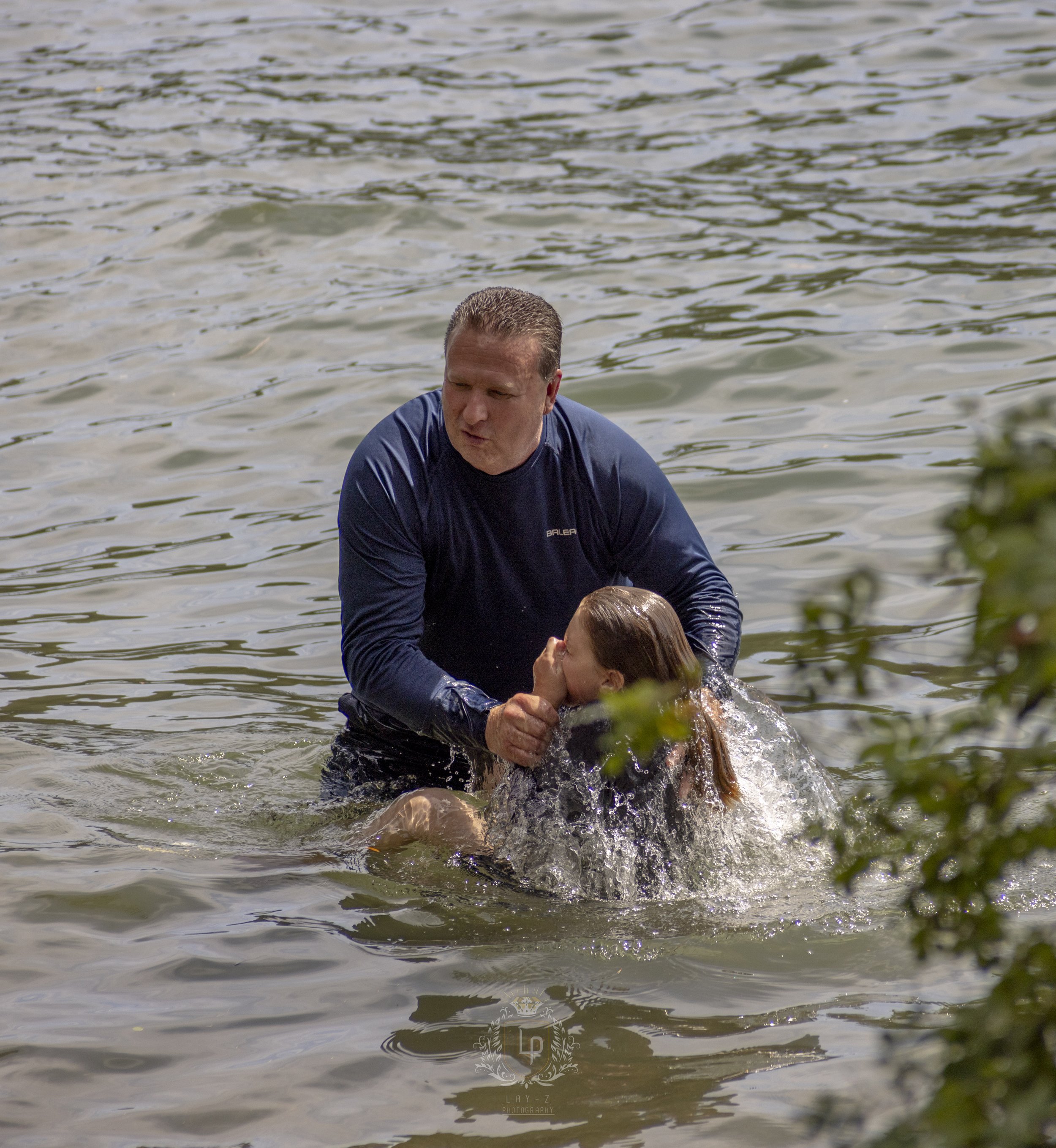A man helping a young girl out of a body of water, with the girl covering her face and the man holding her head, in an outdoor setting with water and some blurred greenery in the foreground.