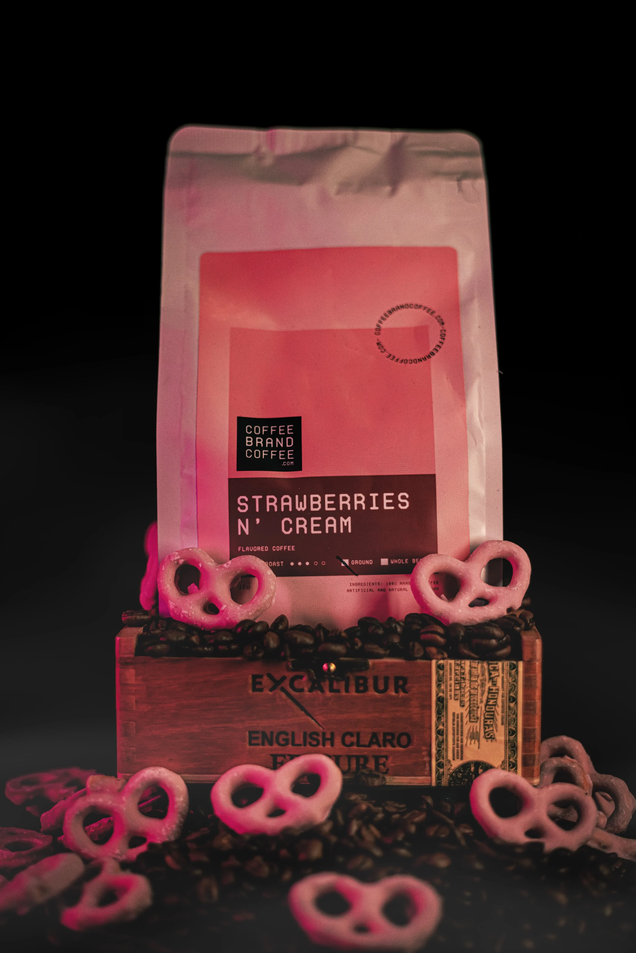 A bag of coffee labeled "Strawberries N' Cream" sitting on a wooden box filled with coffee beans and pink pretzels, with additional pretzels scattered around the base, against a black background.