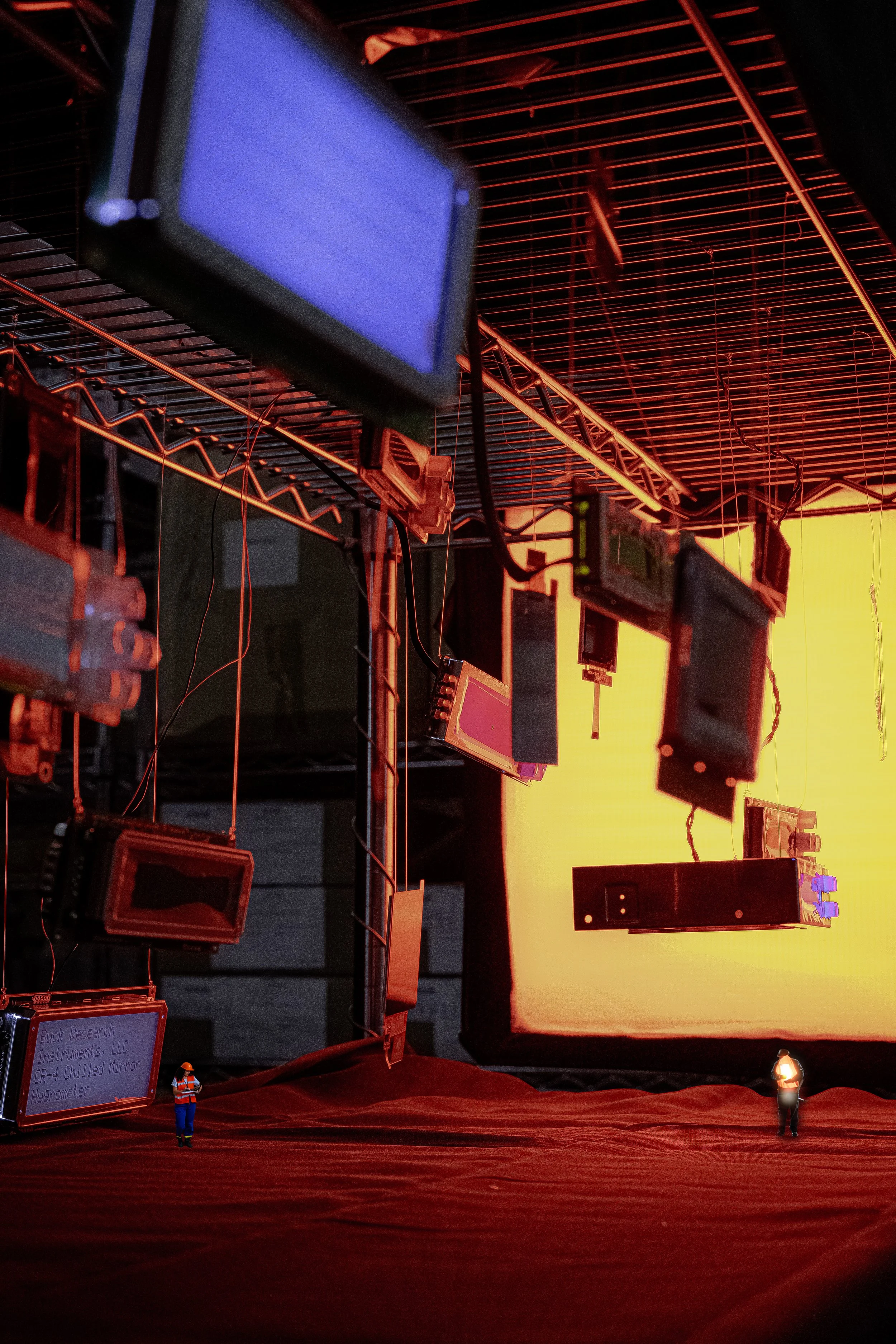 A miniature scene of a production studio with tiny workers in safety vests and helmets, set against a red-lit industrial background with hanging electronic screens and equipment.