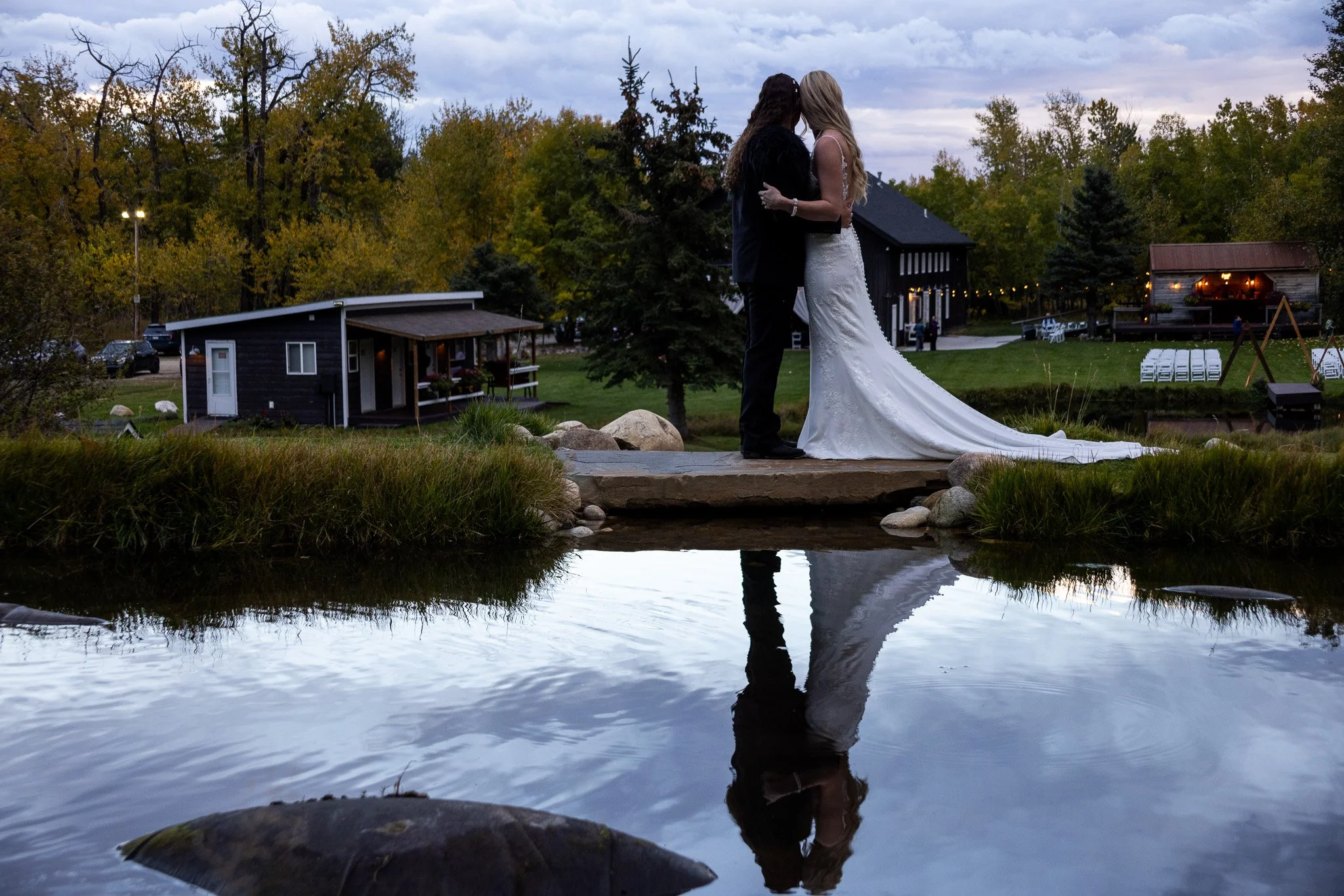 A couple dressed in wedding attire standing on a small stone bridge over a pond, embracing and touching foreheads during sunset.