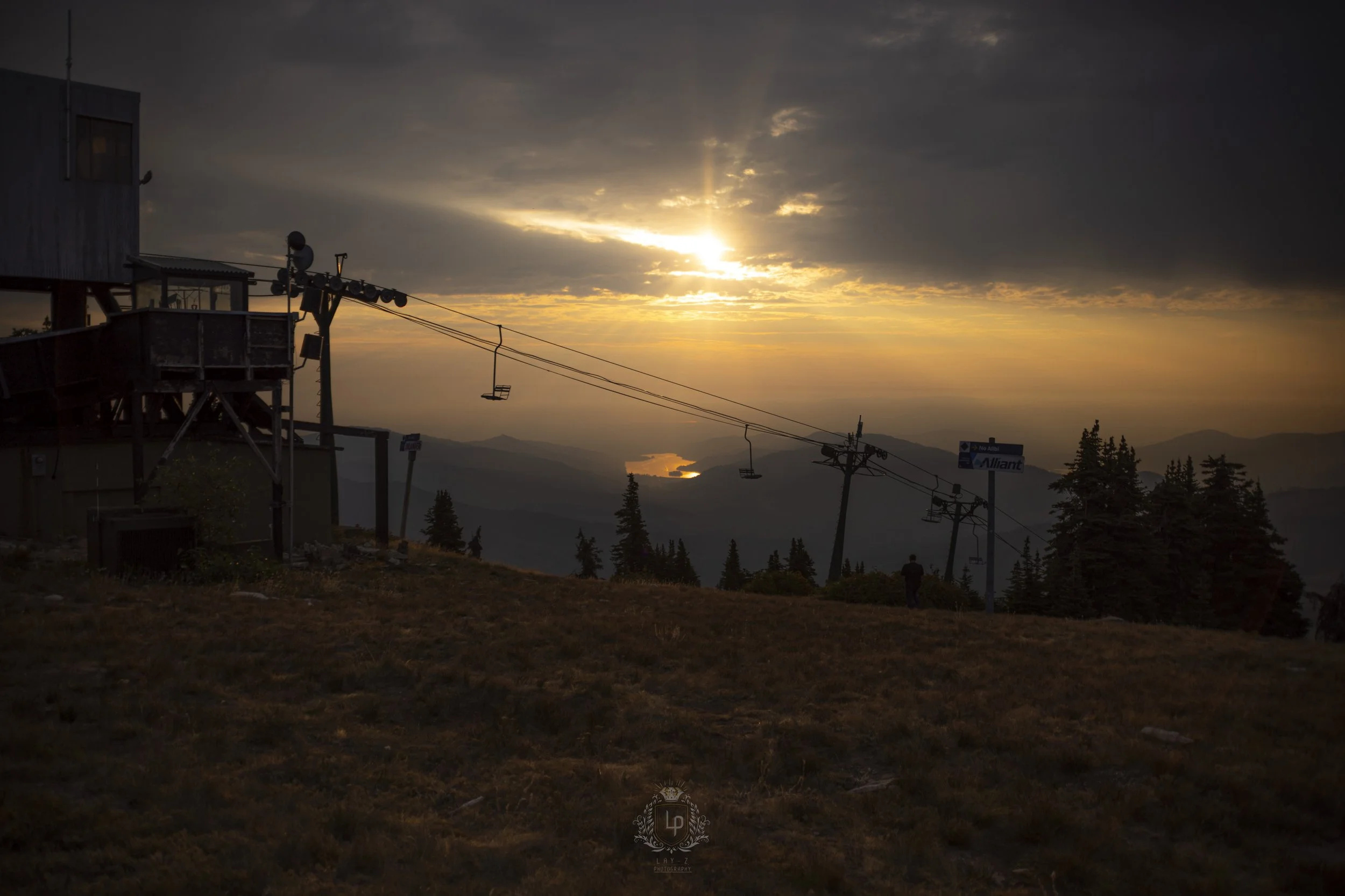 Sunset over mountainous landscape seen from a ski lift area with empty chairs hanging from cables, a weathered building on the left, and trees in the distance.