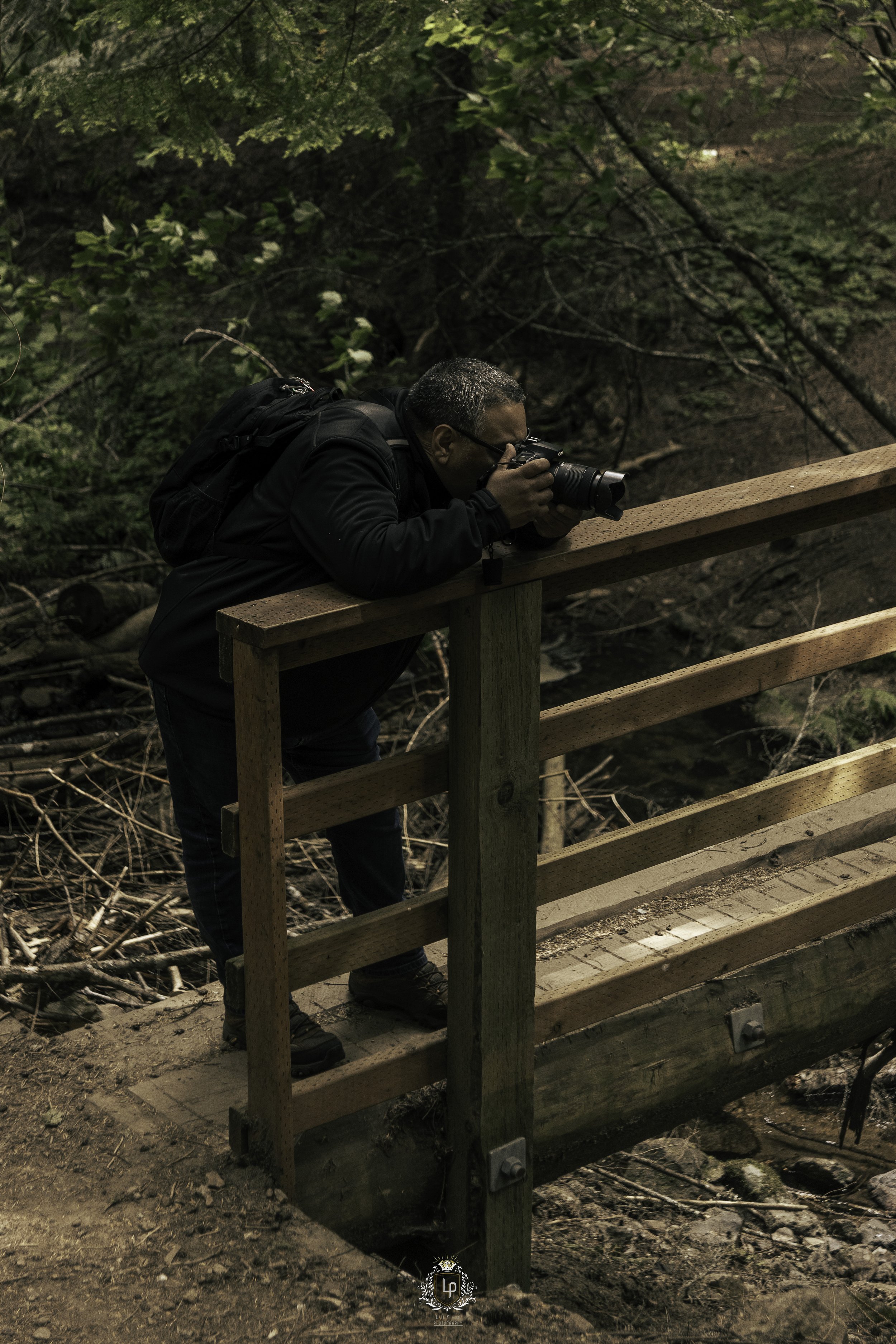 A person with a backpack is taking a photograph with a camera from a wooden bridge in a wooded area.