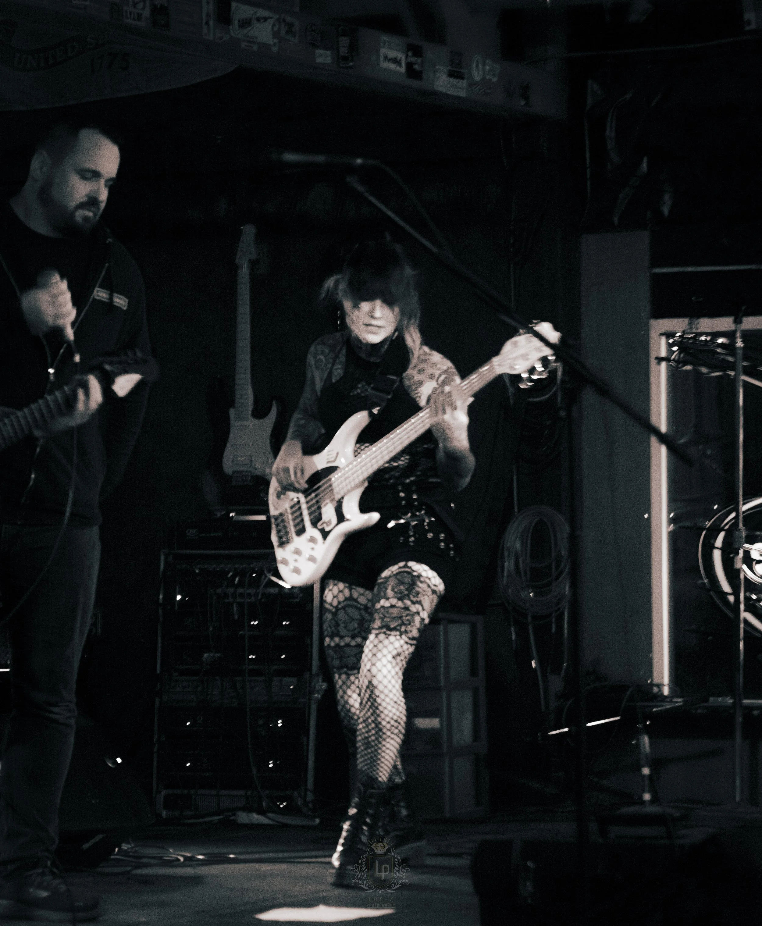 Female musician playing an electric bass guitar on stage during a live performance, with a dark background and male musician with a guitar beside her.