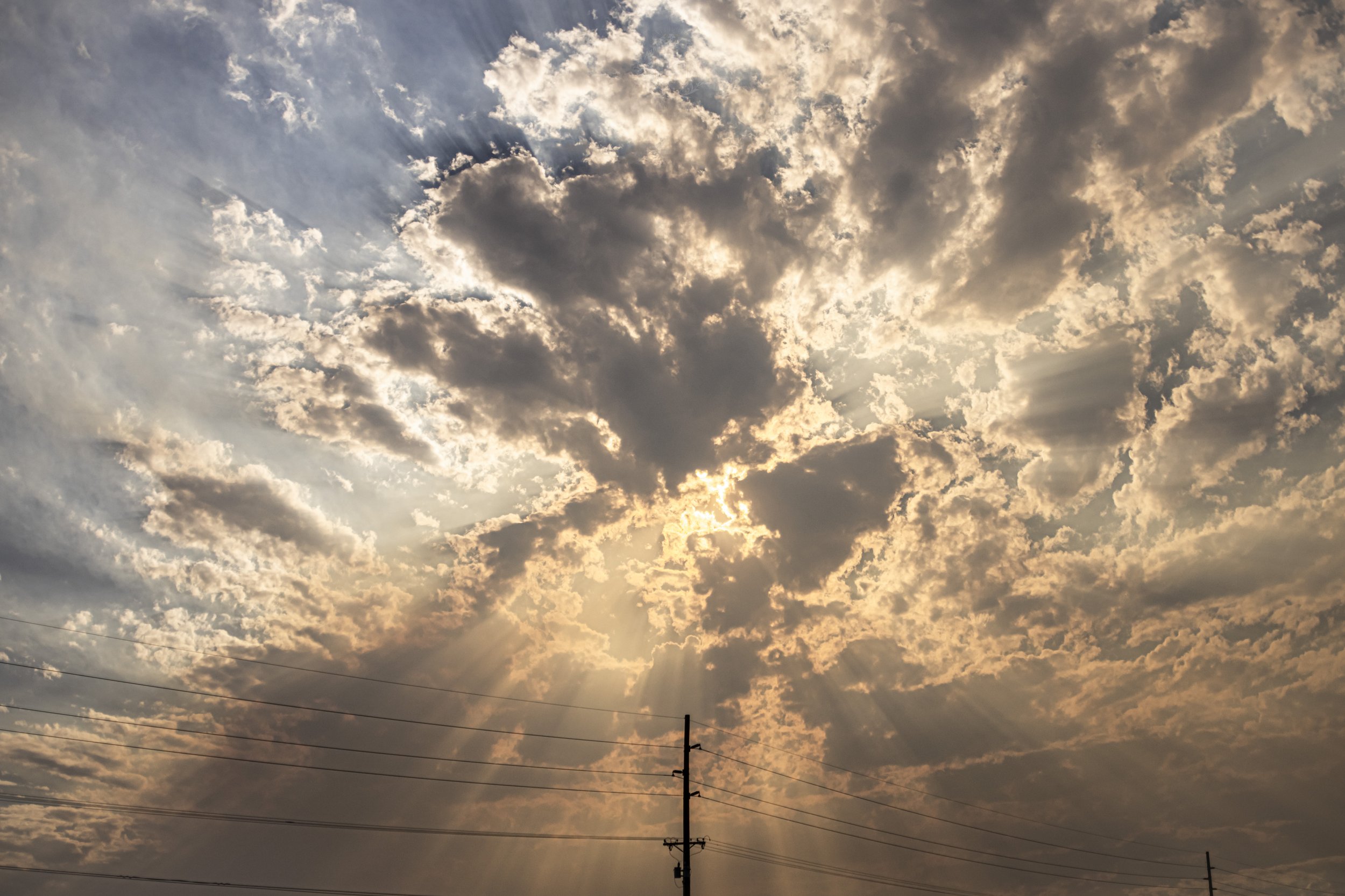 Cloudy sky with the sun partially obscured by clouds, producing sun rays. Power lines and a utility pole are visible in the foreground.