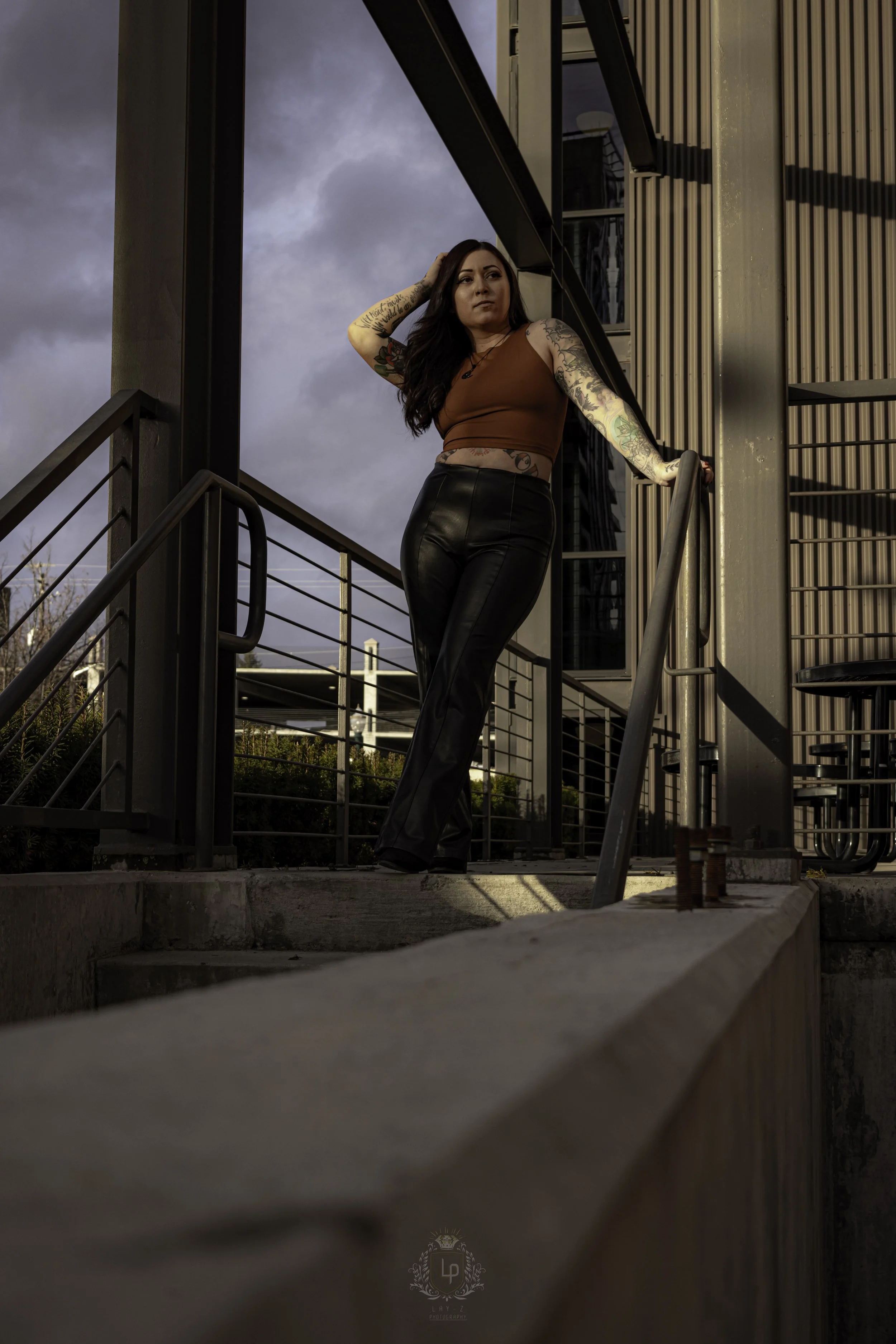 A woman with tattoos, wearing a brown crop top and black leather pants, standing on an outdoor staircase, leaning on a railing, with dark clouds and a modern building in the background.