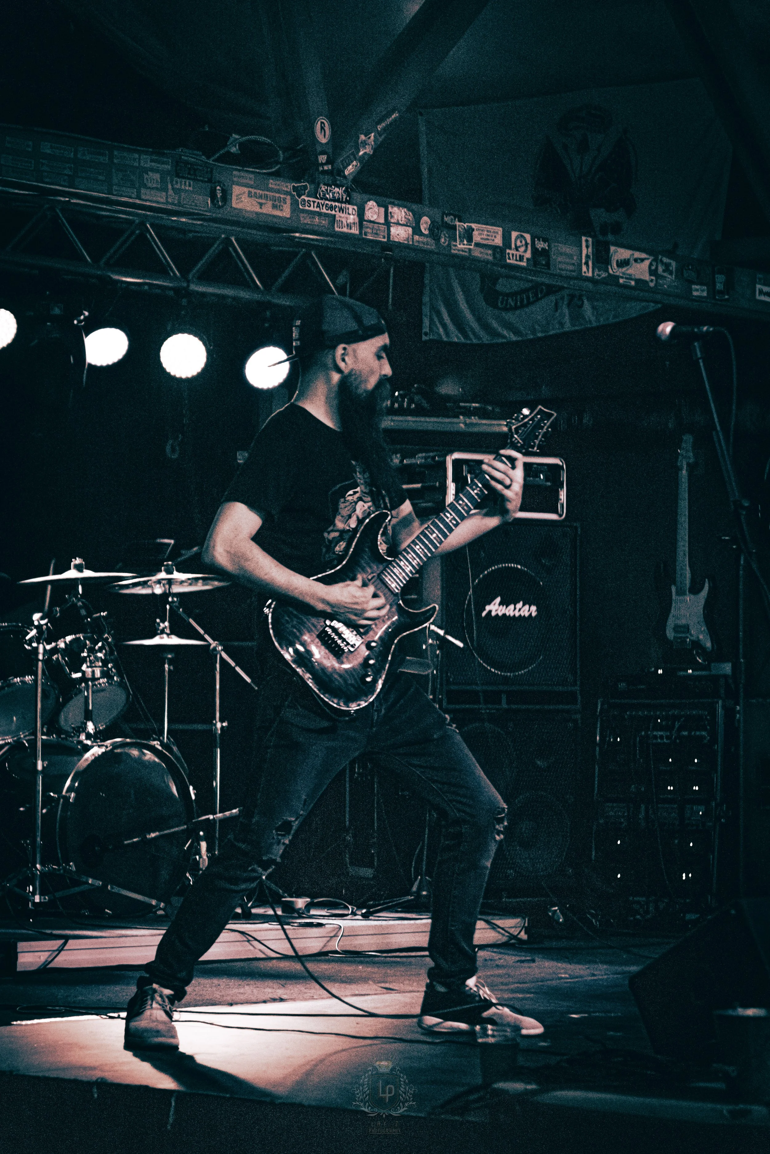 A musician with a beard, wearing a backward cap, black t-shirt, and ripped jeans, playing an electric guitar on stage during a live performance. There is a drum set behind him and various amplifiers and equipment around him.