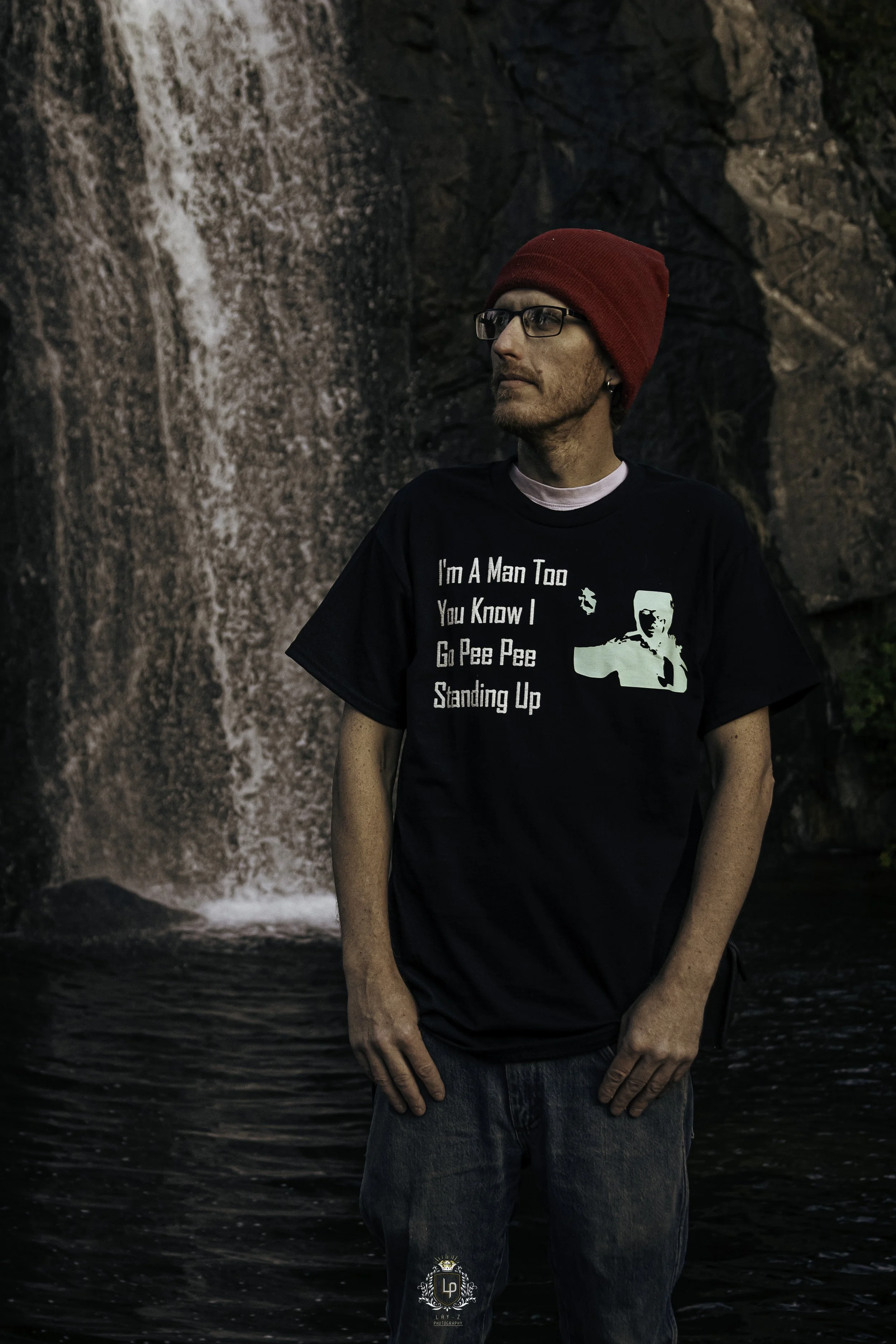 A man wearing a red beanie, glasses, and a black T-shirt with white text stands in front of a waterfall with rocks and green foliage in the background.