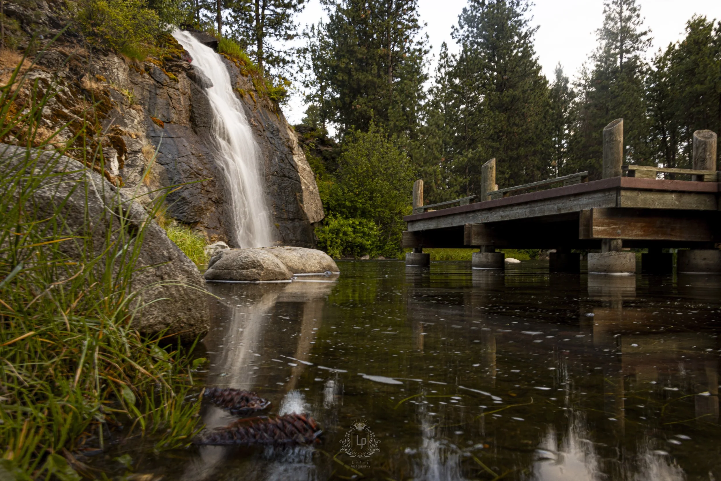 A peaceful scene of a waterfall flowing over rocks into a calm pool, with a wooden footbridge over the water and a forest of pine trees in the background.