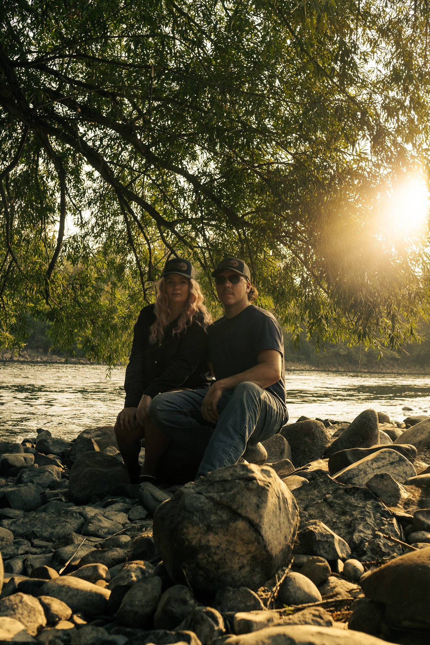 A couple sitting on rocks by a river during sunset, with large leafy trees overhead.