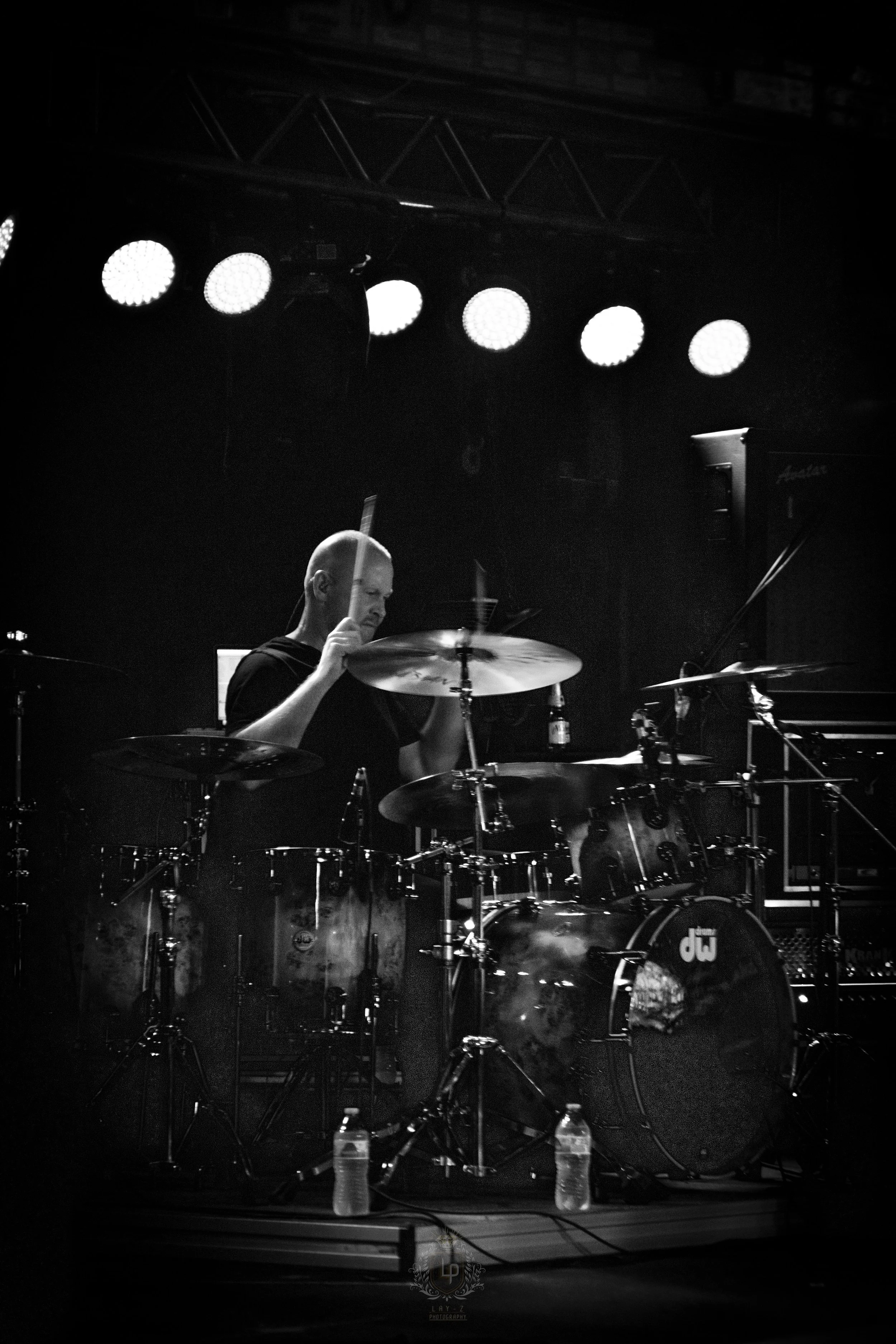 A black-and-white photo of a drummer playing a drum set on stage, under bright stage lights.