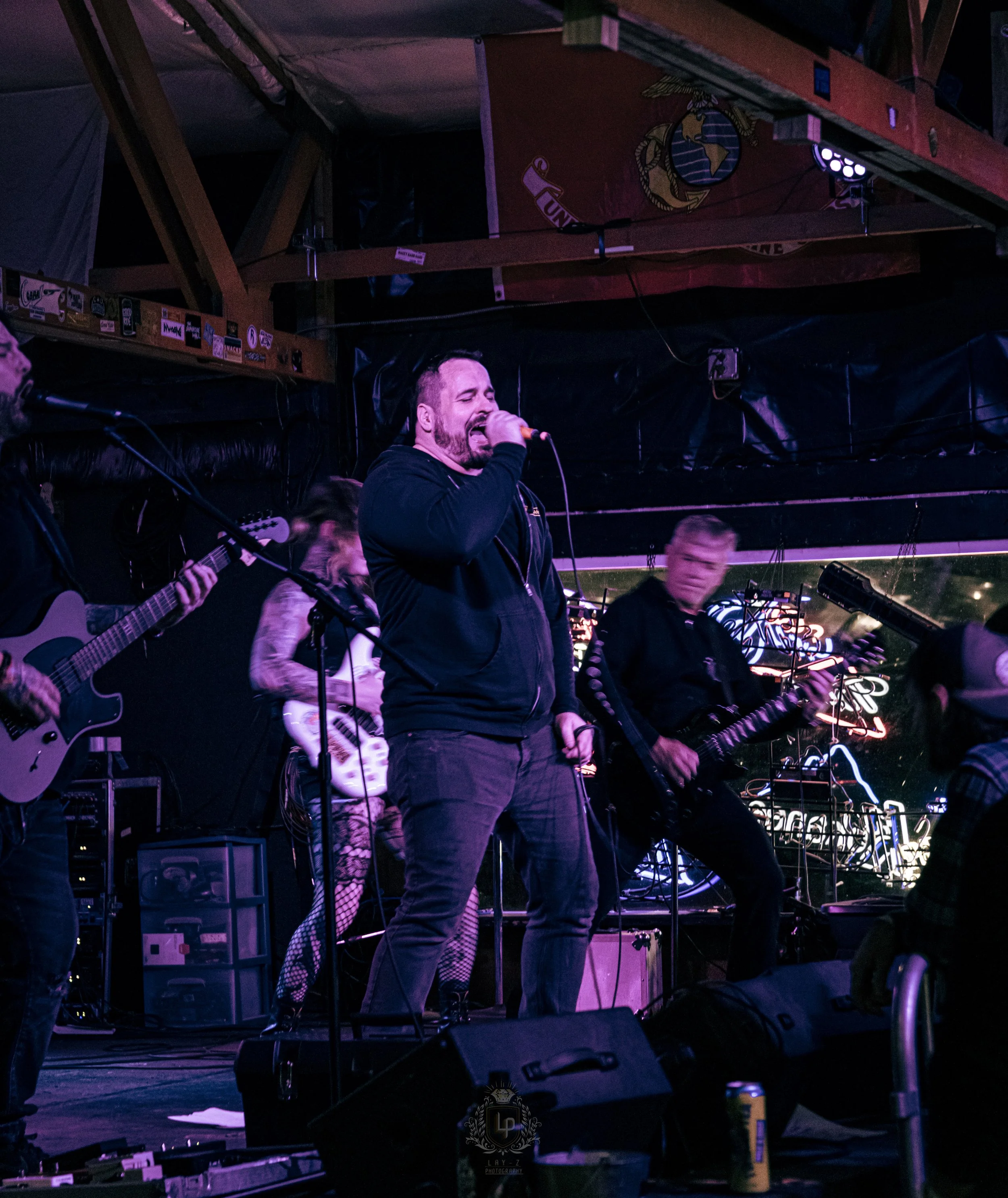 A band performing on stage with a man singing into a microphone, surrounded by guitar players, in a dimly lit venue with colorful lighting and a neon sign in the background.