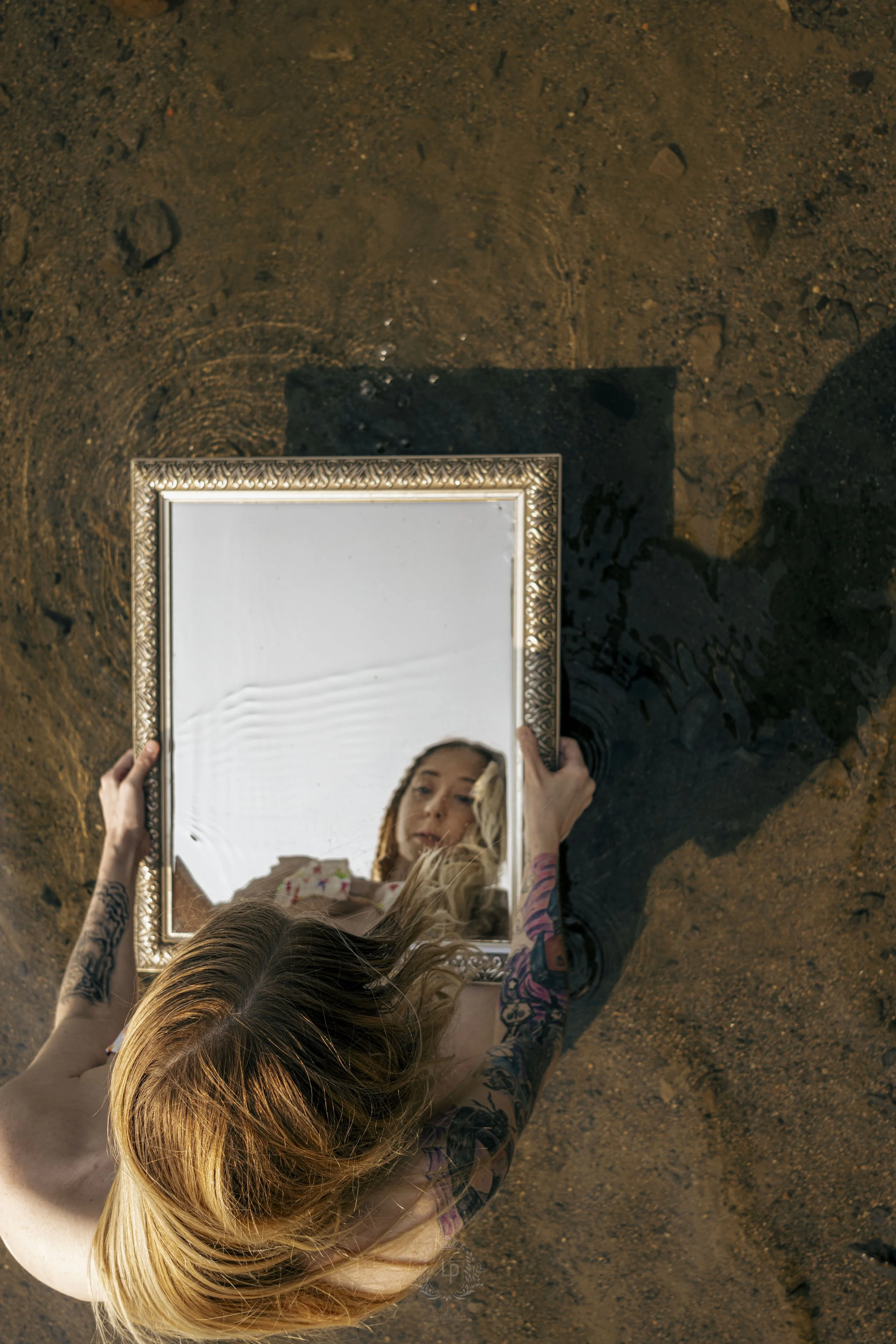 A woman with tattoos on her arms is sitting on sandy ground, holding a decorative mirror that reflects her face, with her blonde hair falling over her shoulders.