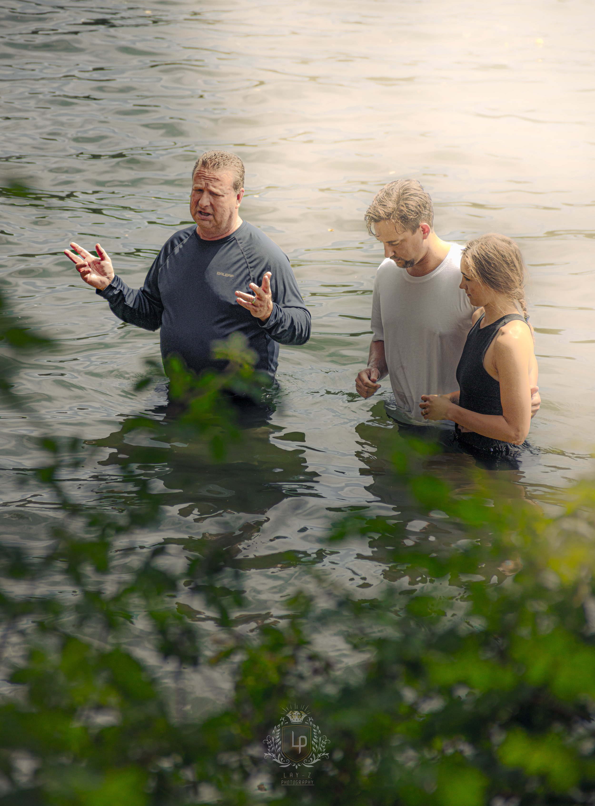 Three people standing in a body of water, possibly a lake or river. One man is speaking and gesturing with his hands, while a man and woman listen. Green foliage partially obscures the foreground.