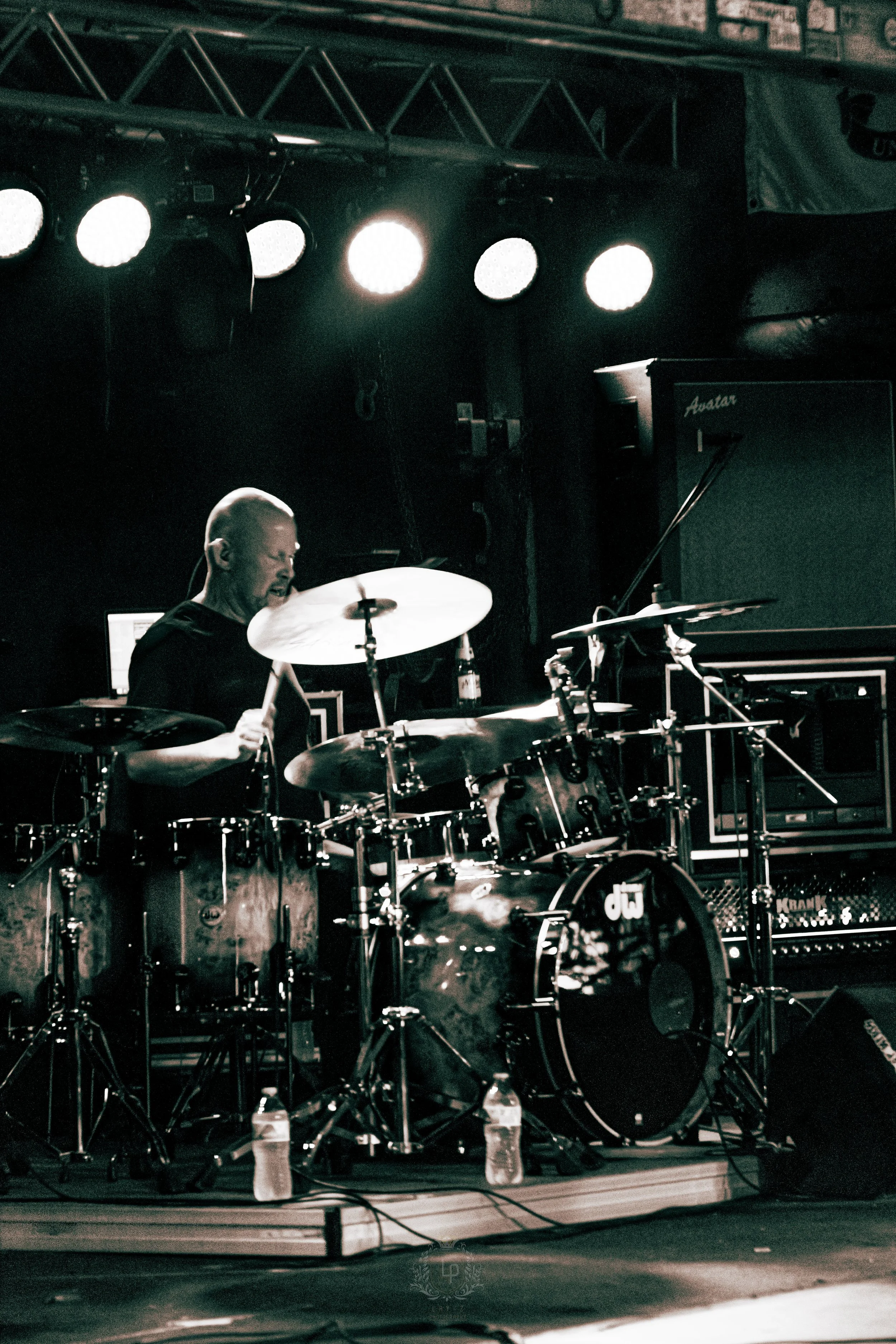 A drummer playing on stage with bright stage lights overhead, a drum set, and music equipment in the background.