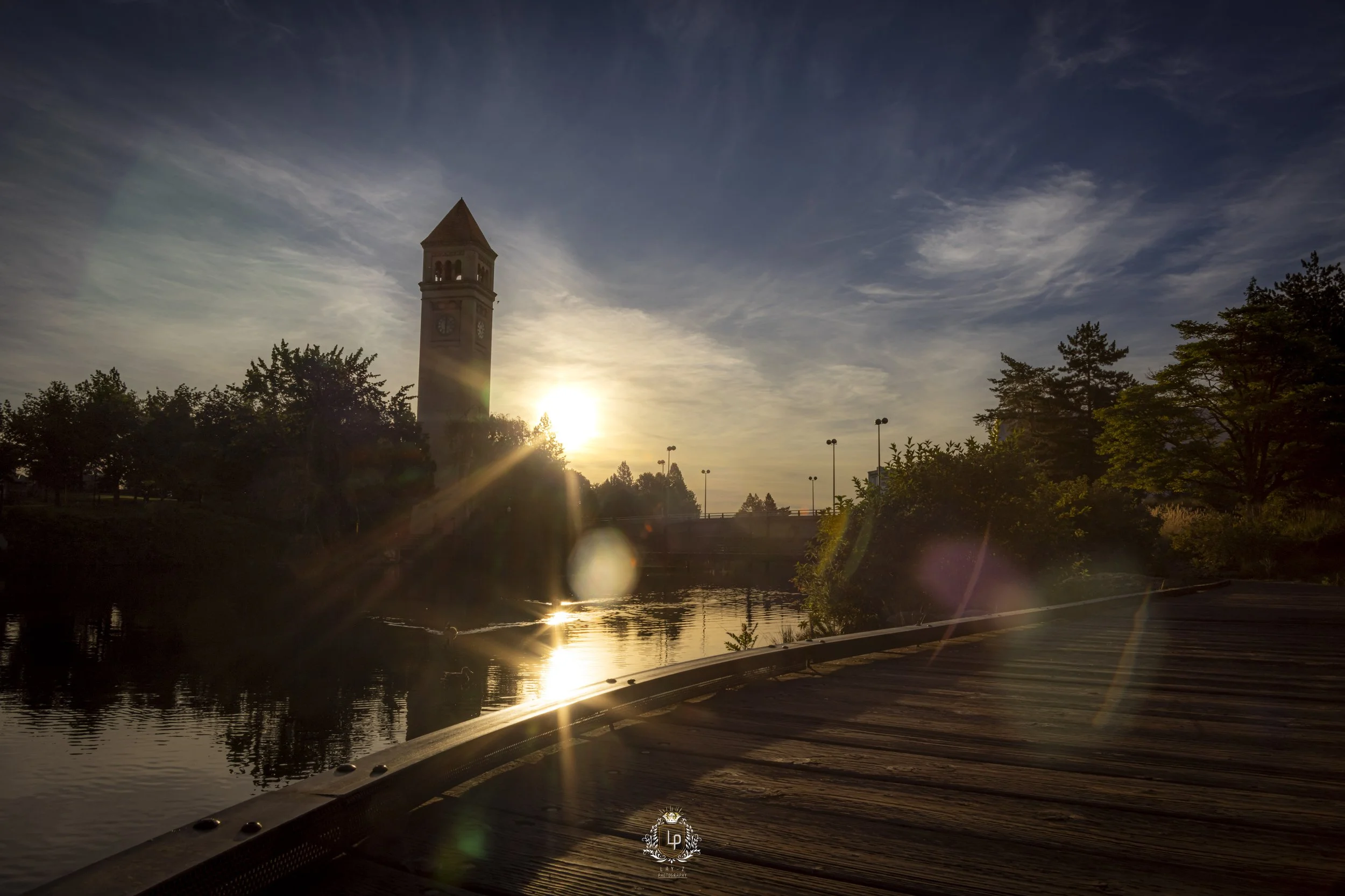 Sunset over a clock tower by a river, with trees and a wooden bridge in the foreground.