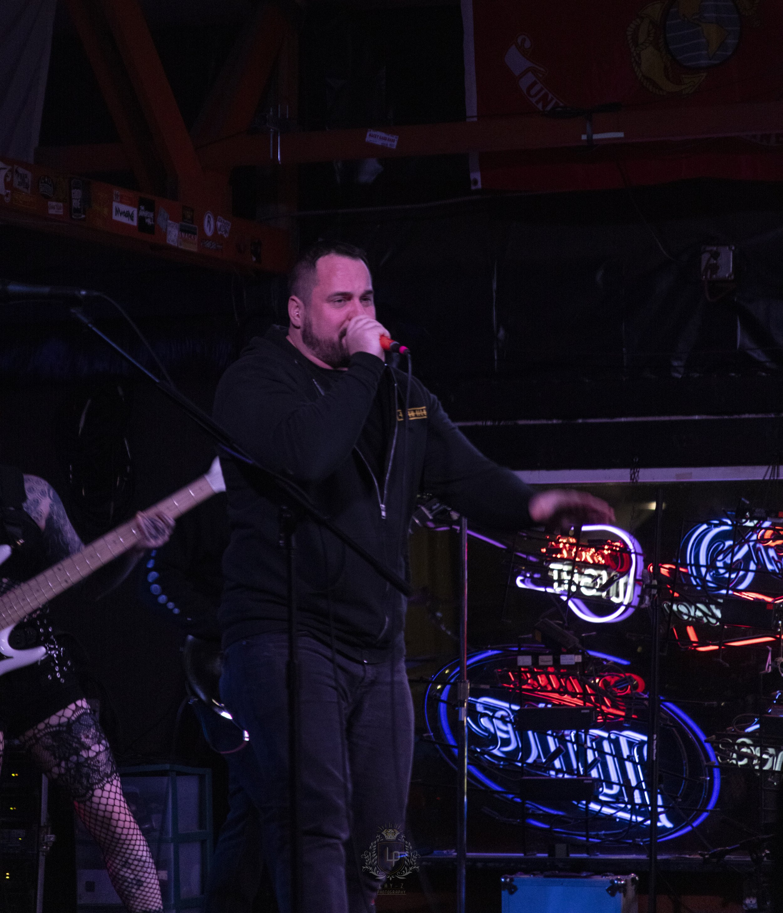 A man singing into a microphone on stage with neon signs in the background.