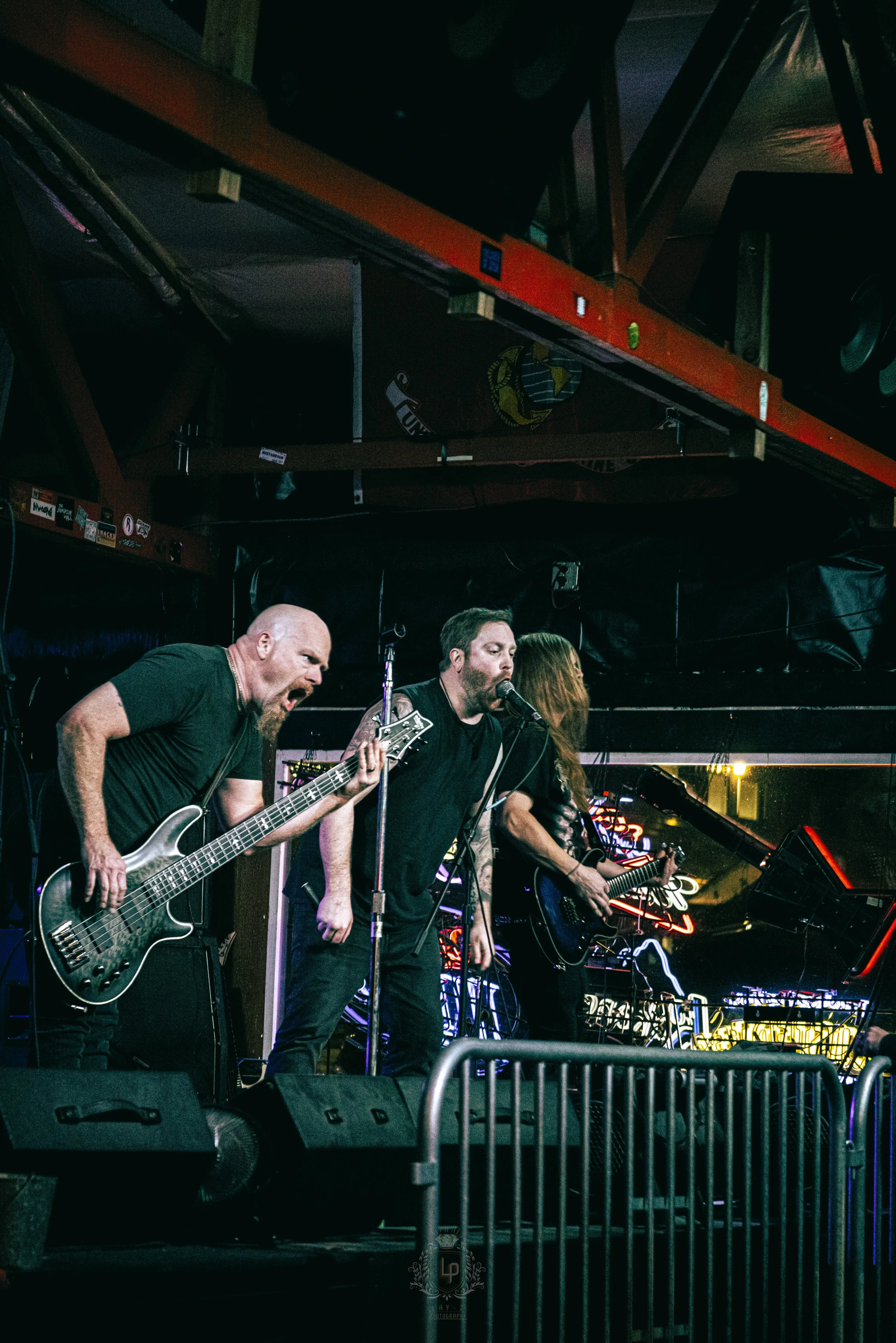 Three musicians performing on stage at a concert venue, with one guitarist passionately playing and singing, another guitarist slightly behind, and a bassist to the side, surrounded by colorful neon lights.