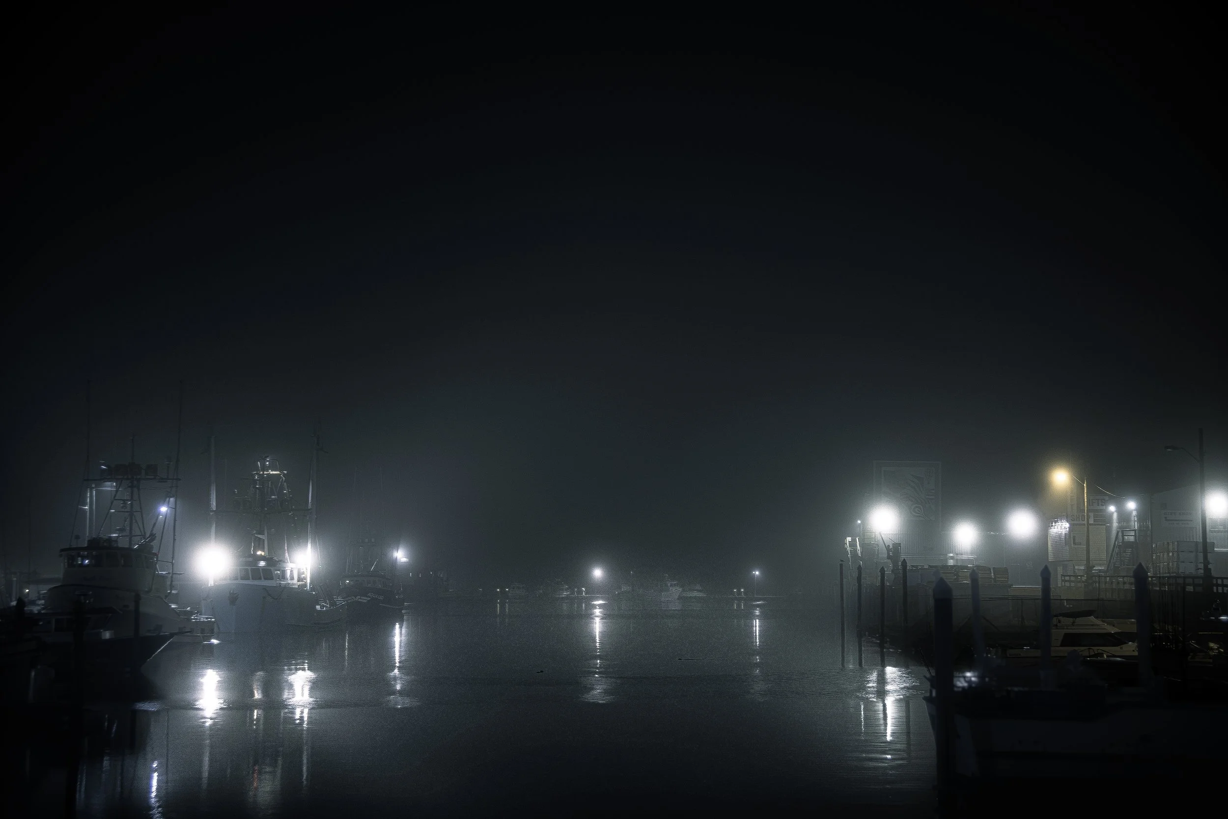 Nighttime scene of a foggy marina with boats docked, lit by bright white lights, reflected on the calm water.