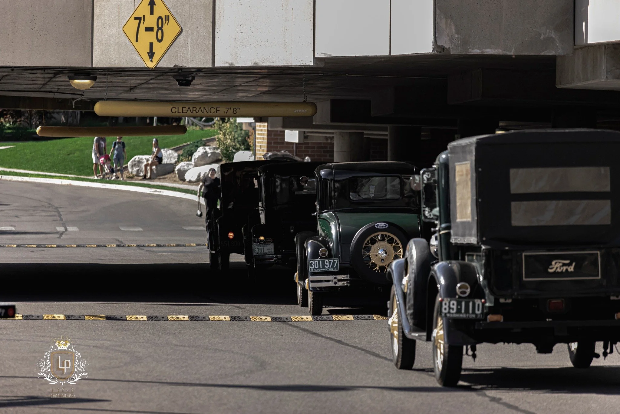 Vintage black cars parked under a building overhang with a clearance of 7 feet 8 inches, with people and a green grassy area in the background.