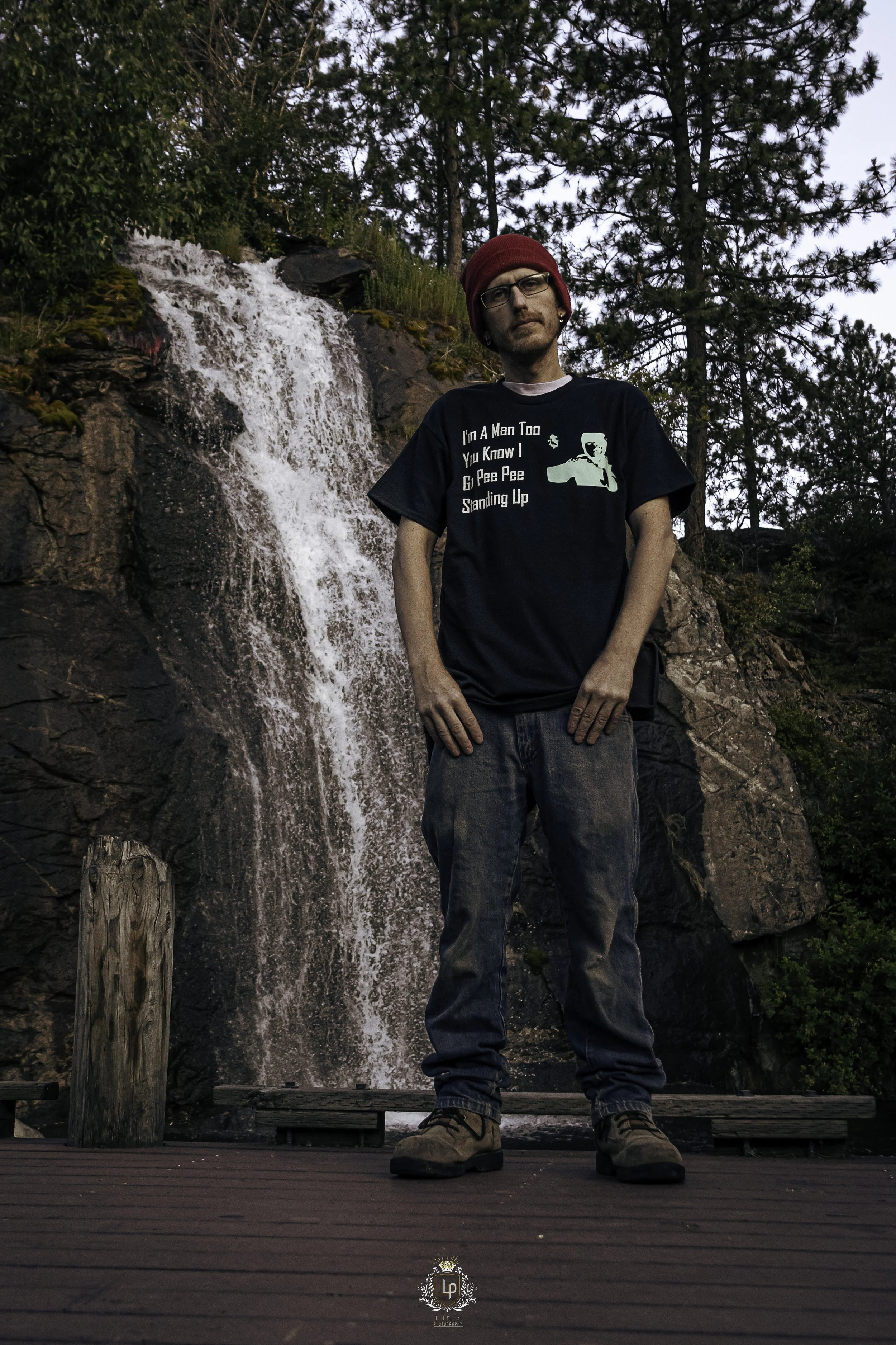 Person standing on a wooden platform in front of a waterfall surrounded by trees, wearing a red beanie, glasses, a black t-shirt with white text and a graphic, and jeans.