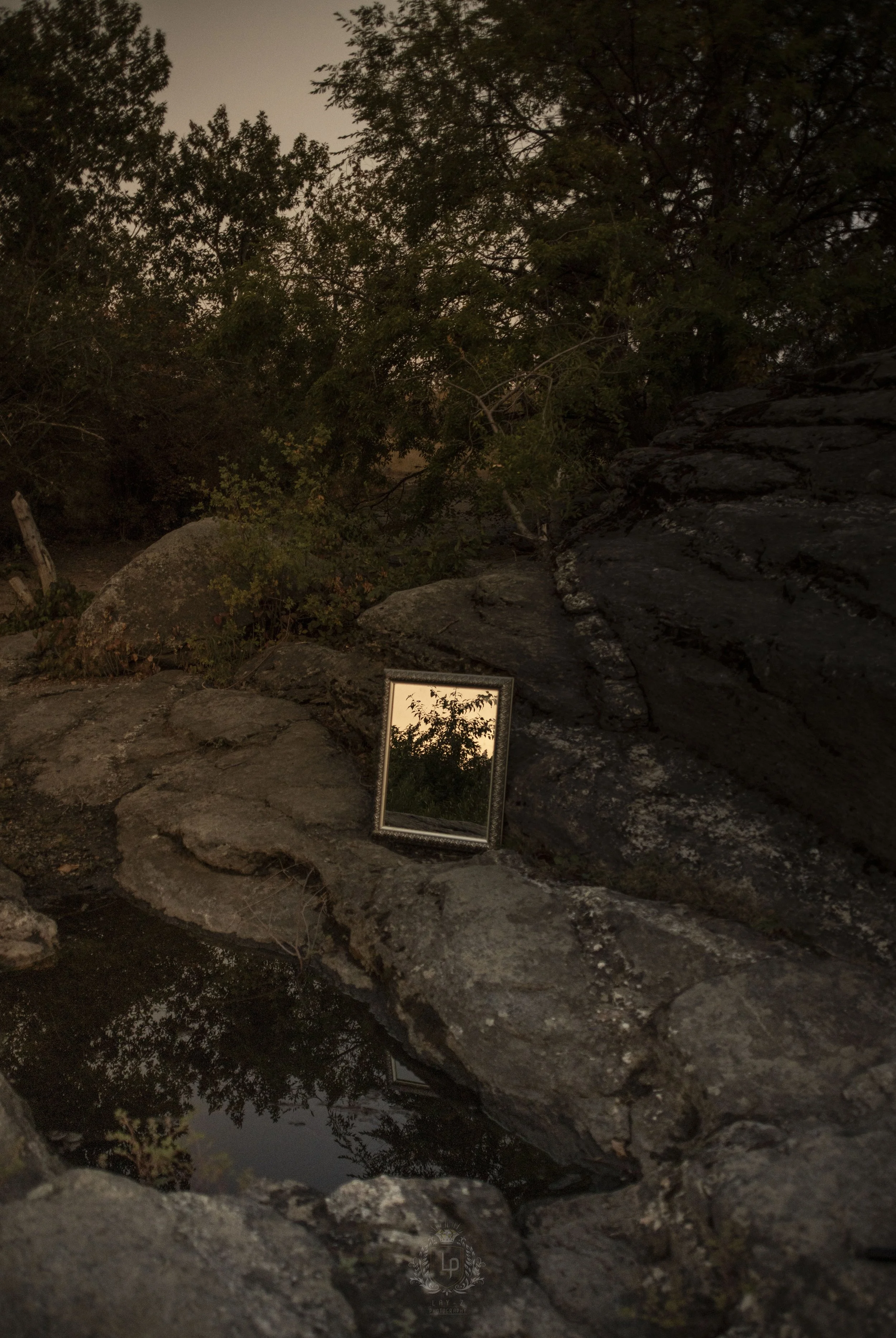 A small rectangular mirror leaning against rocks outdoors, reflecting trees and the sky at sunset or dusk, surrounded by large rocks and trees with fading light.