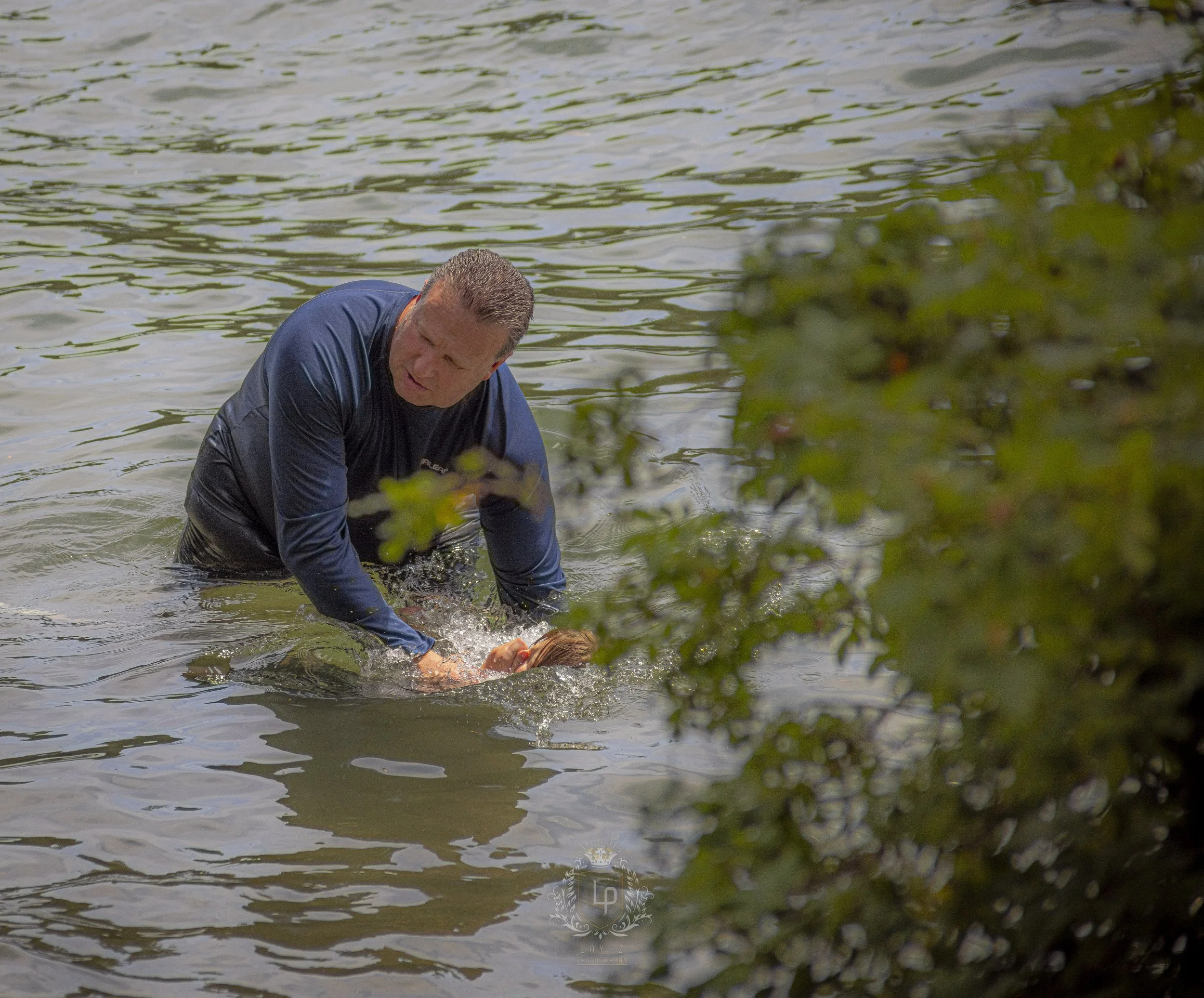 A man in a navy shirt supports a person with red hair in the water during a rescue or training exercise near a bush.