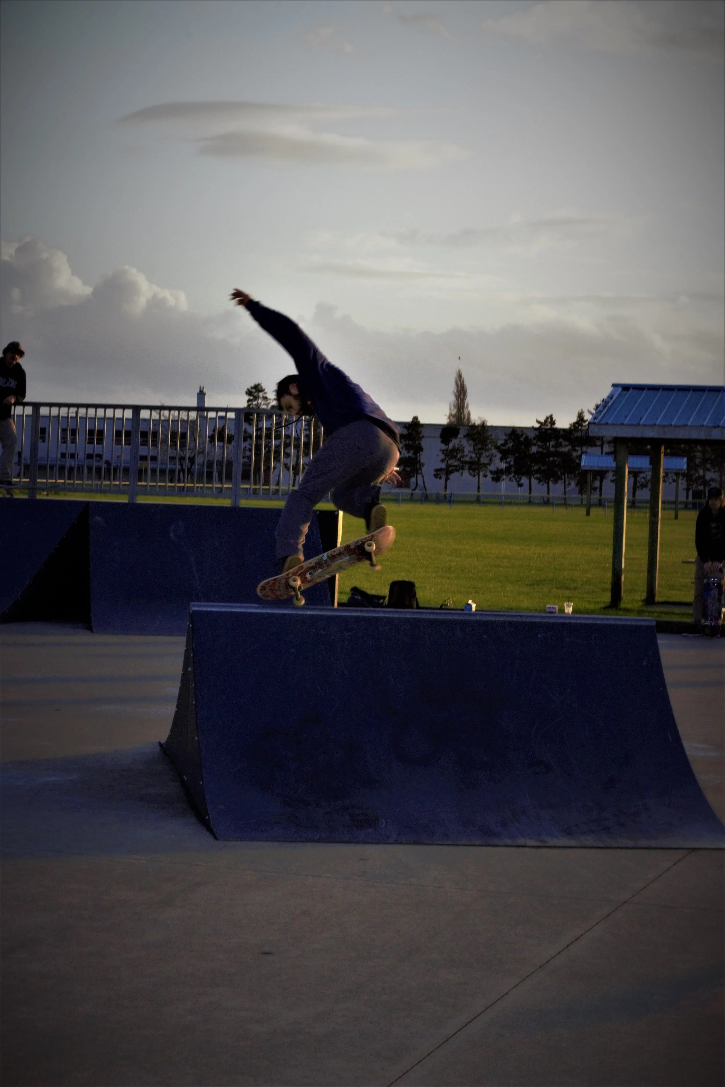 Skateboarder performing an ollie trick at a skatepark during the late afternoon or early evening, with a woman sitting under a shaded structure in the background and a cloudy sky overhead.