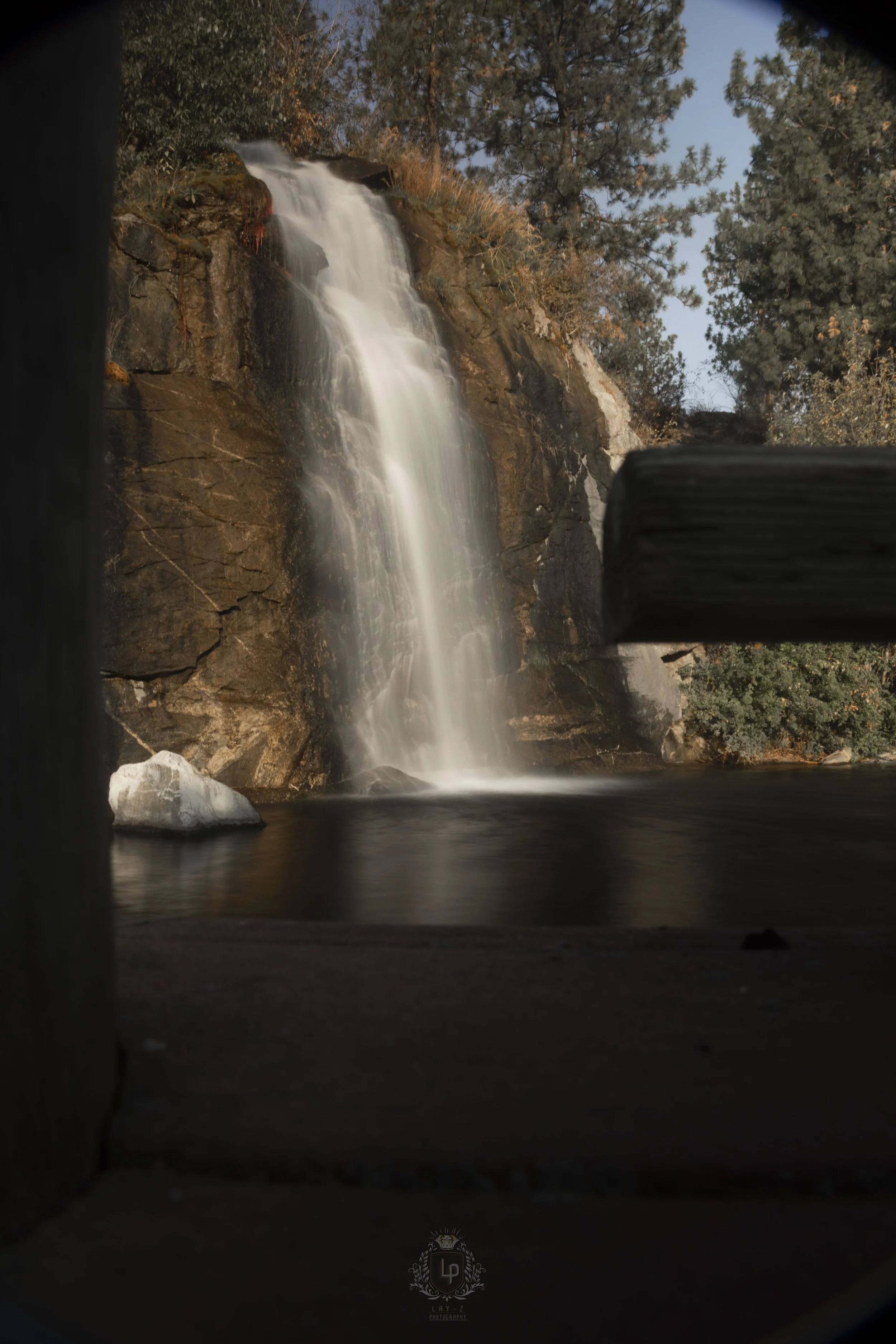 A waterfall flowing over rocks into a pond, framed by wooden posts, surrounded by trees in a natural setting.
