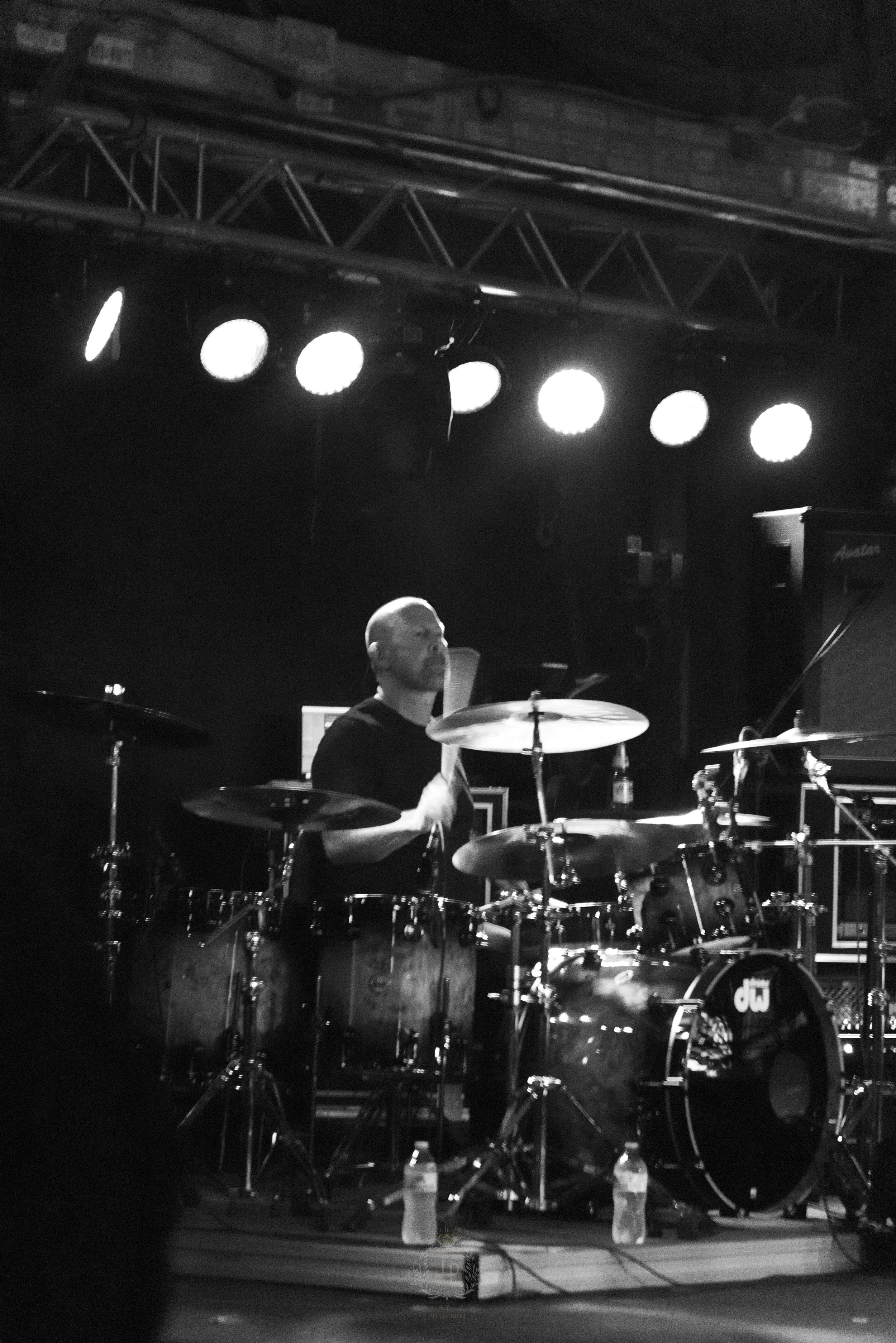 A drummer playing a drum set on stage with bright stage lights overhead.