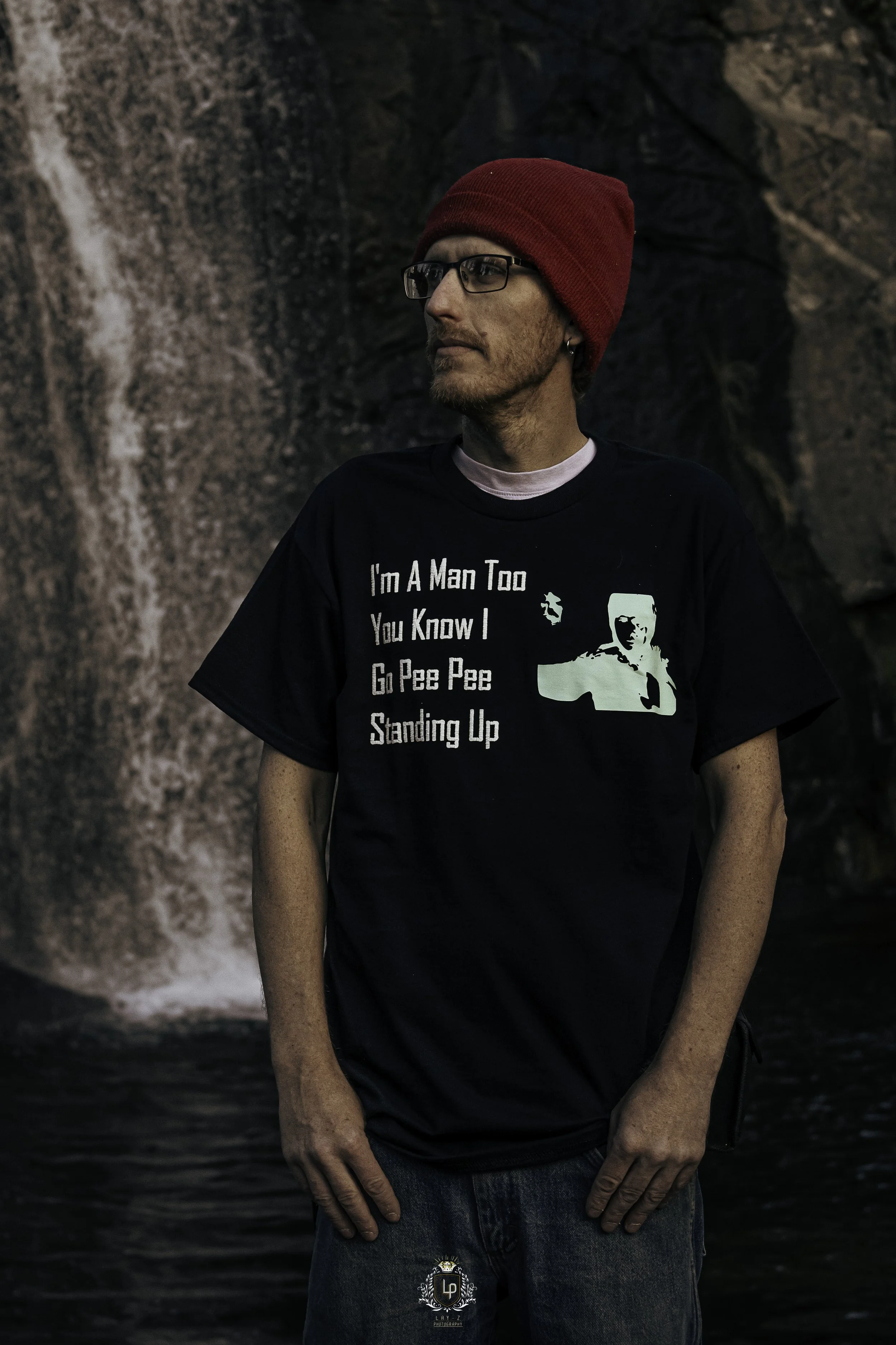 A young man with glasses and a red beanie standing in front of a waterfall. He's wearing a black T-shirt with a humorous message and graphic, and blue jeans.