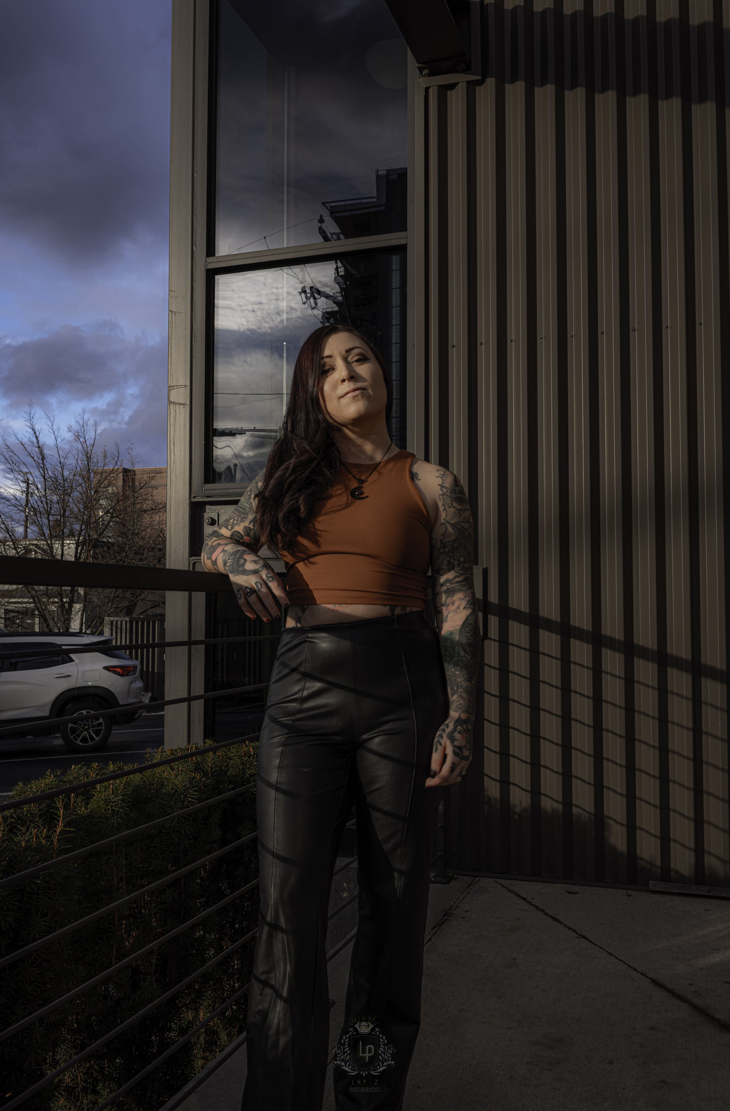 A woman with tattoos on her arms and dark hair, wearing a brown crop top and black leather pants, standing outside on a balcony or terrace with a building and cloudy sky in the background.