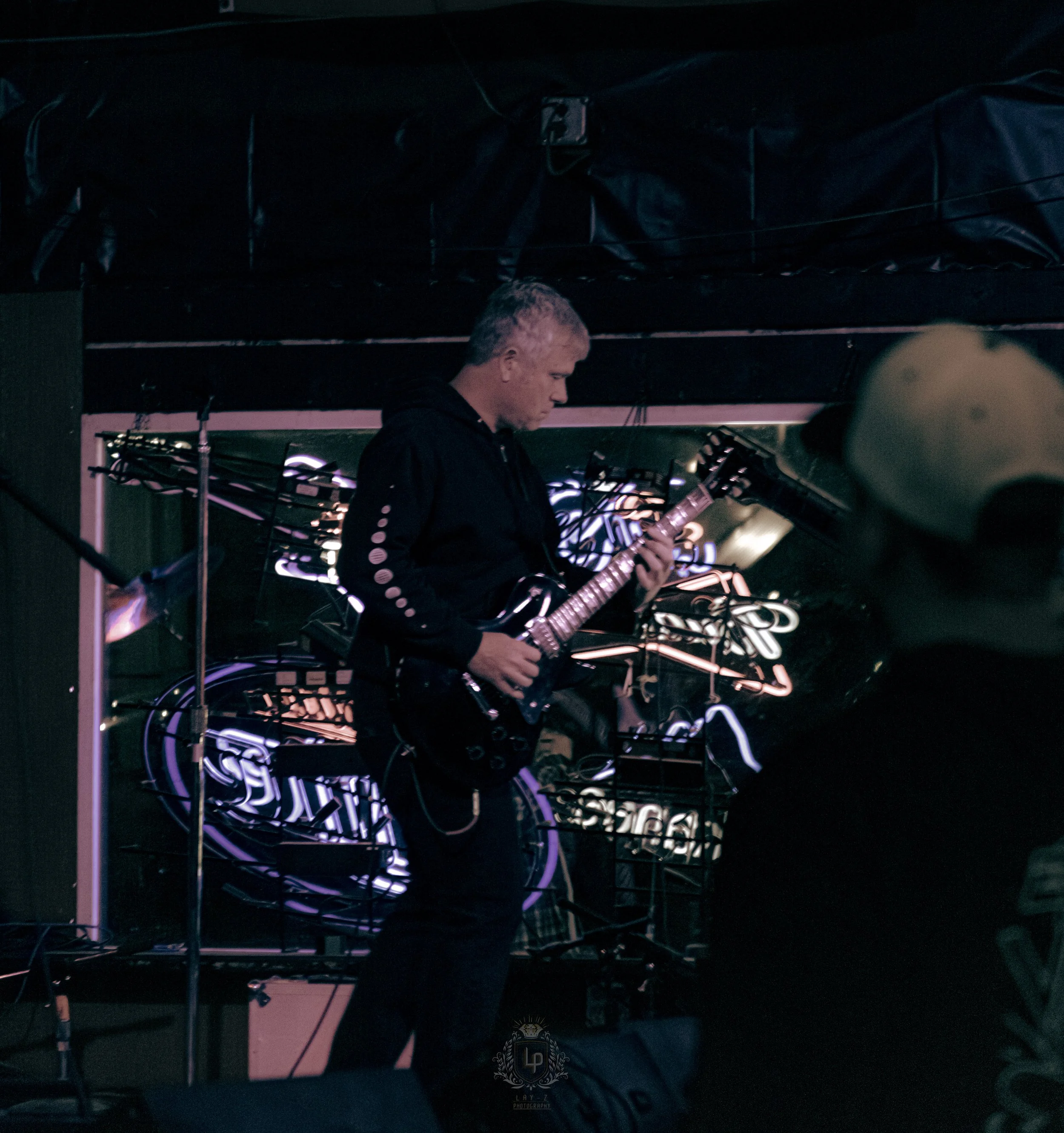 A man playing an electric guitar on a dimly lit stage with neon signs in the background.